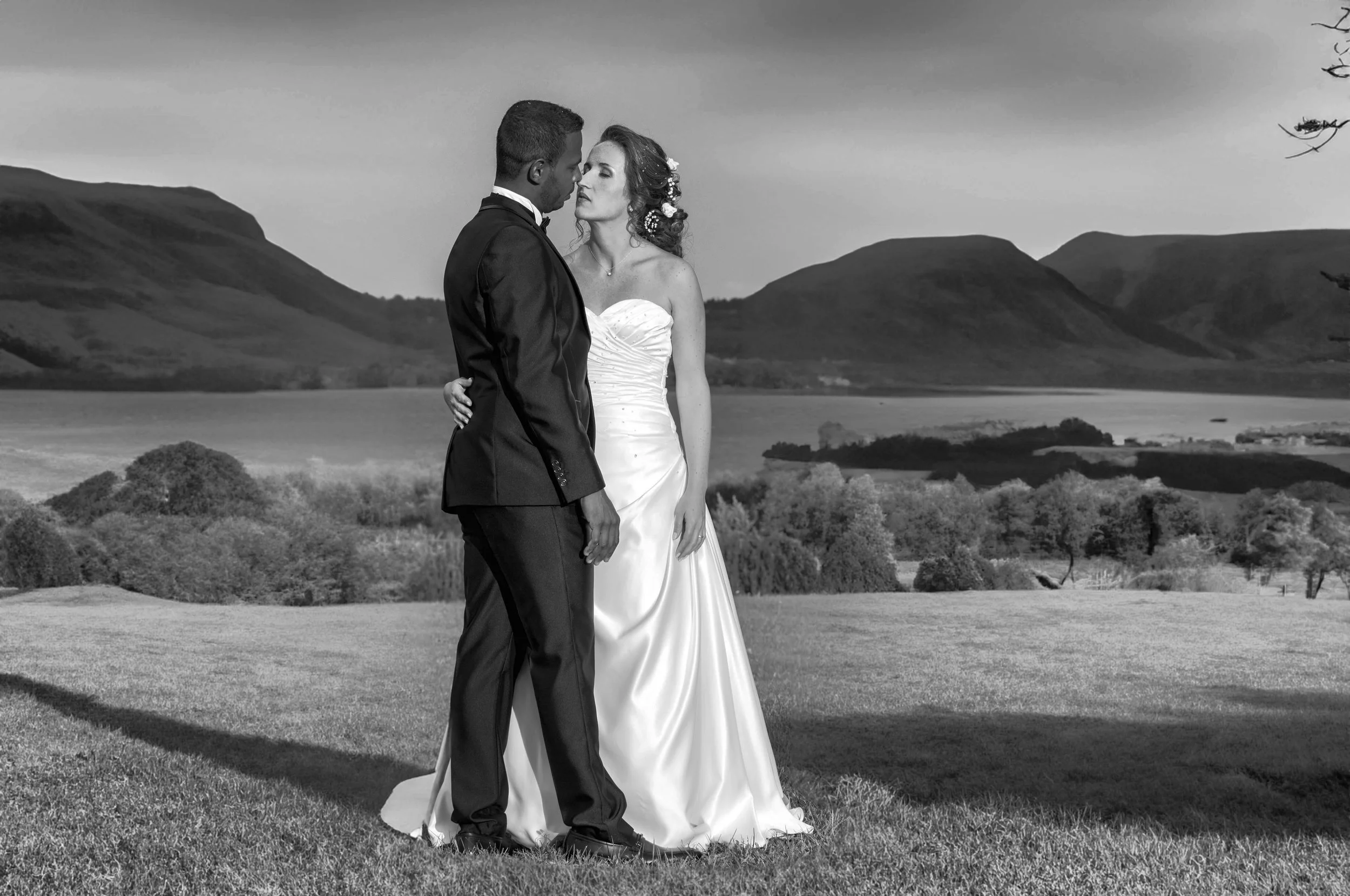 A black-and-white photo of a bride and groom standing close together outdoors with hills and a lake in the background, dressed in wedding attire.