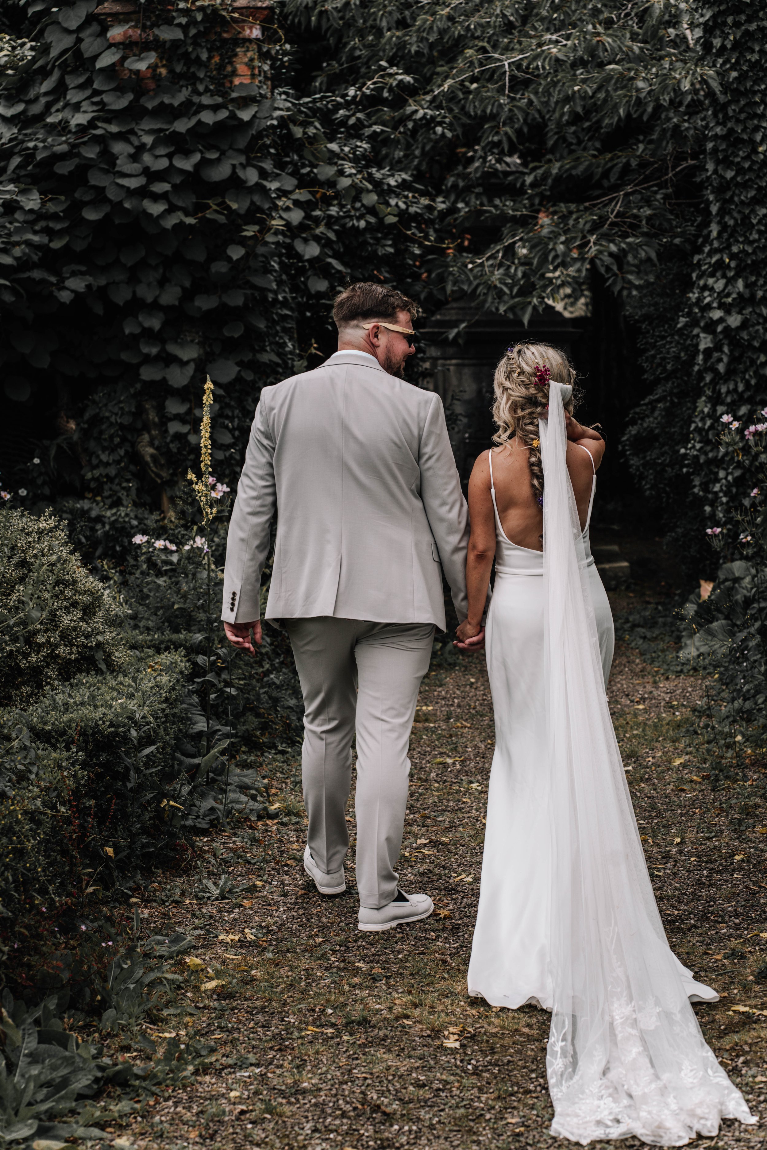 A bride and groom holding hands walking down a garden path, surrounded by lush greenery, on their wedding day.