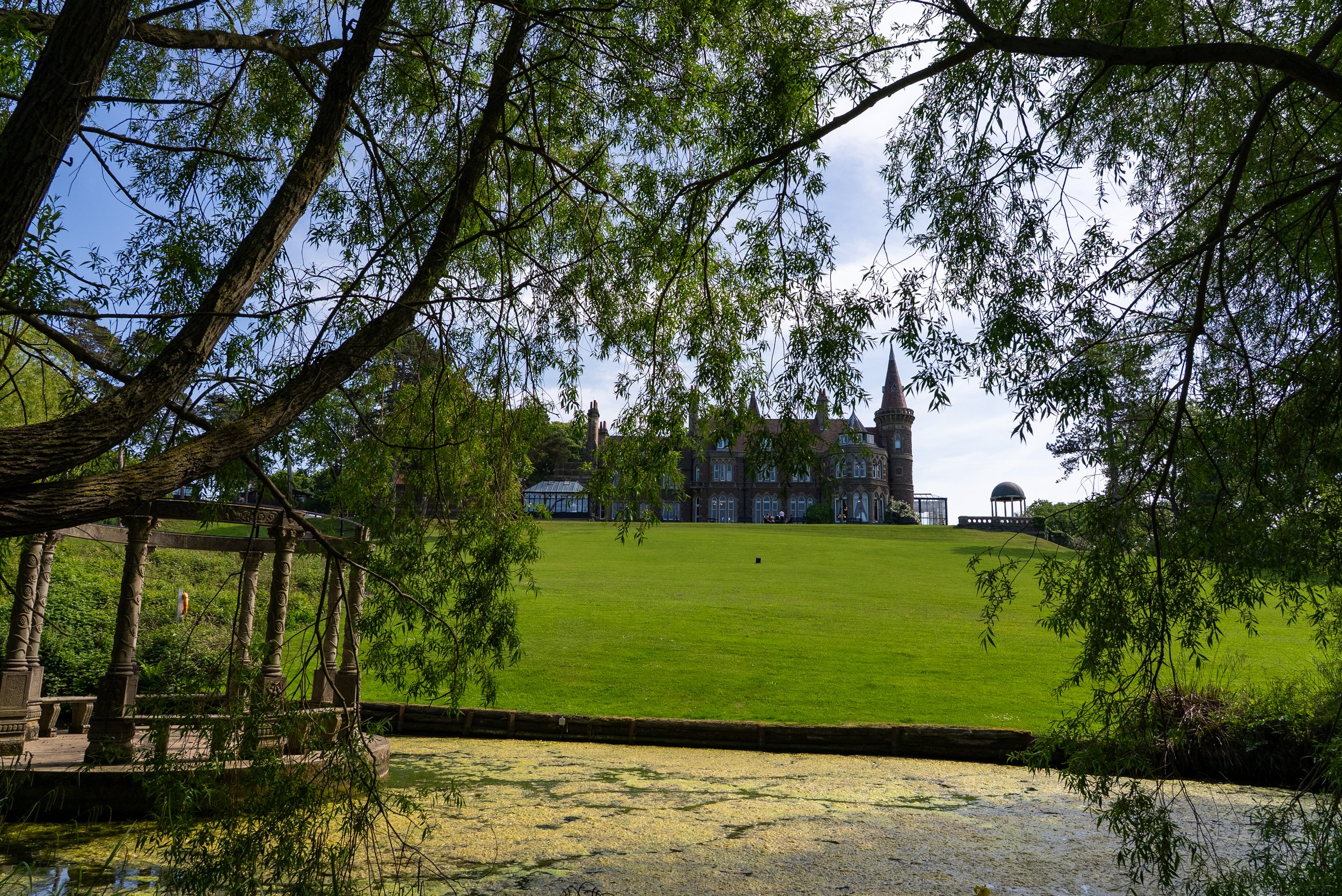 A scenic view of a castle-like house with a tower, framed by tree branches and leaves, overlooking a pond with lily pads and surrounded by lush greenery.