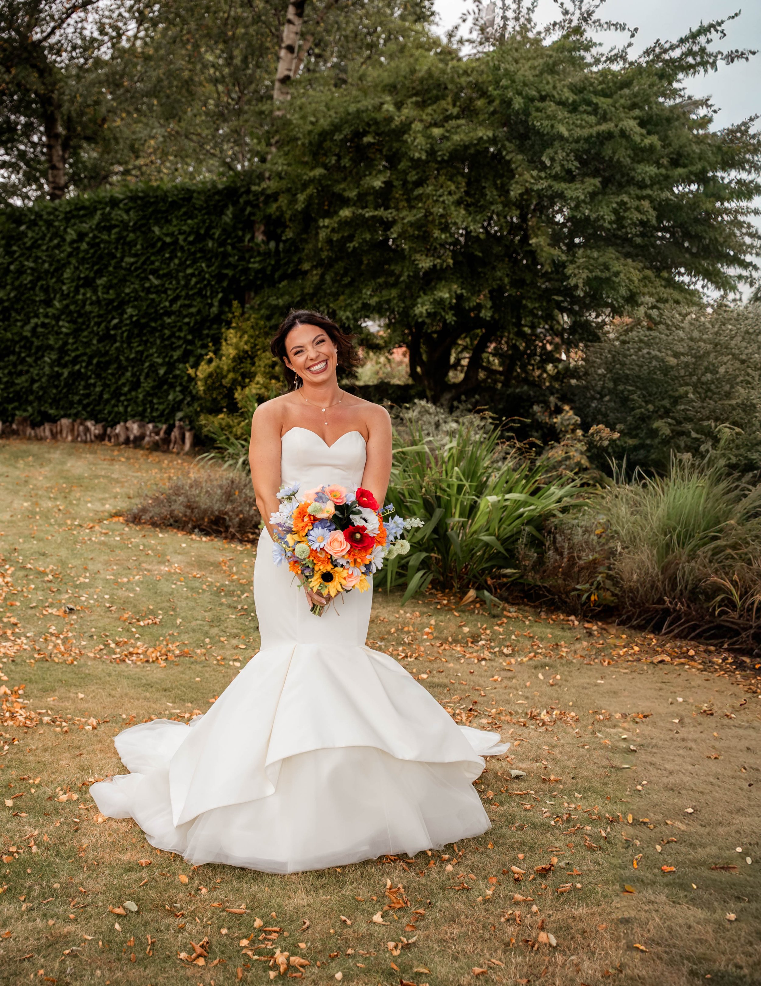 A smiling woman in a white wedding gown holding a colorful bouquet of flowers standing outdoors on a lawn with trees and bushes in the background.