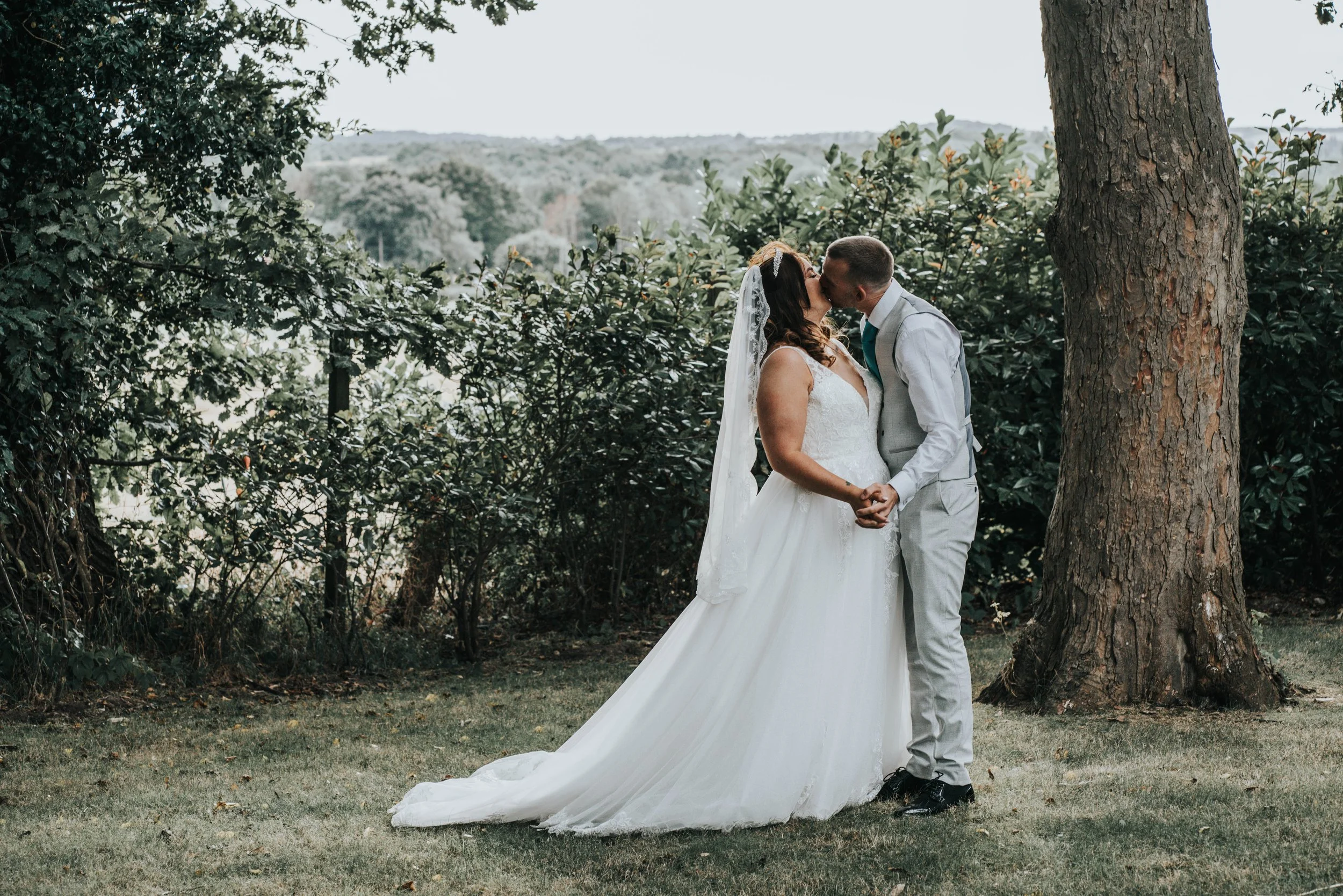A newlywed couple sharing a kiss outdoors next to a large tree, with greenery and a distant hillside in the background.