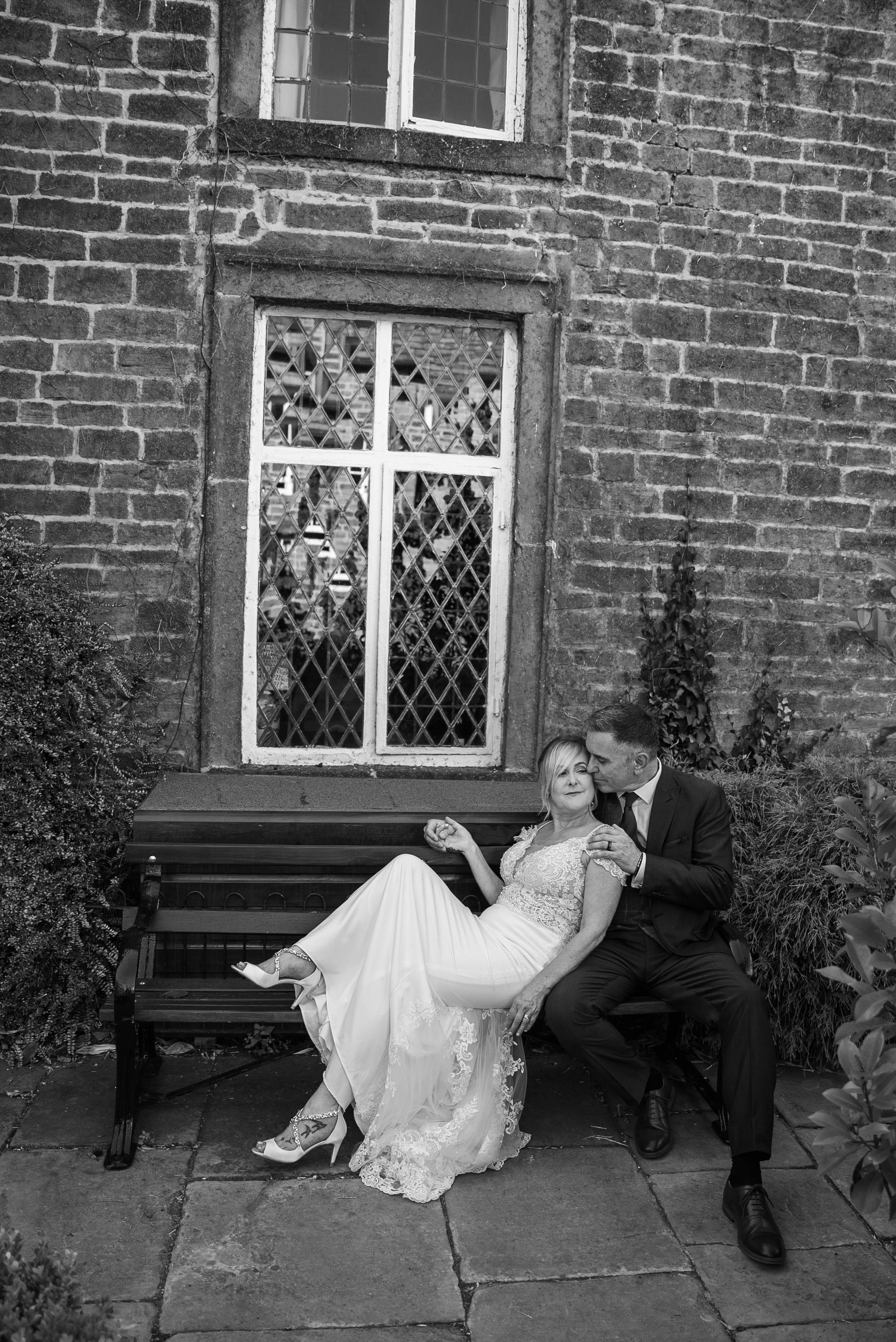 A bride in a lace wedding dress and heels sitting on a park bench with a groom in a black suit and tie, kissing her cheek, in front of a brick building with large multi-pane window.