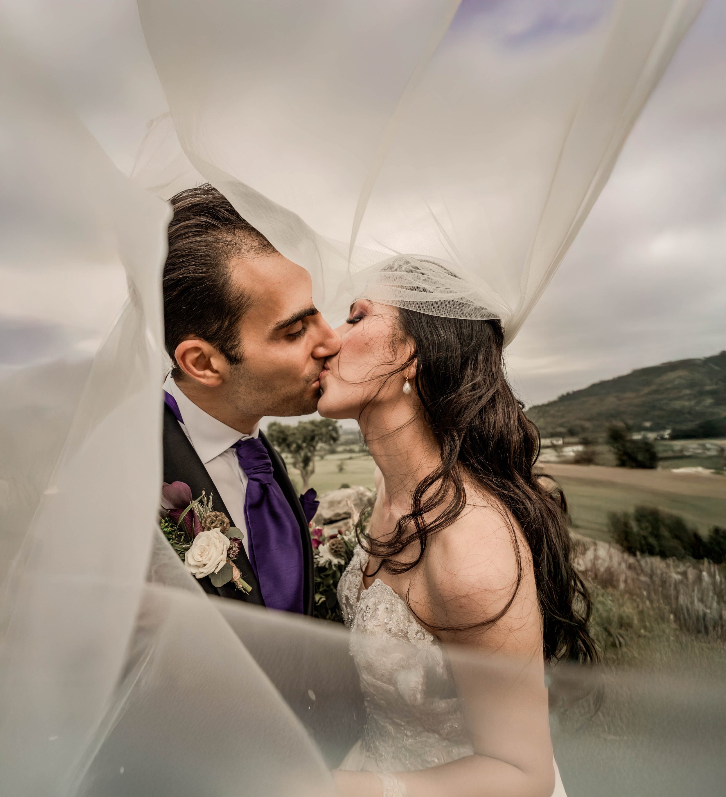 A bride and groom kissing outdoors under a sheer wedding veil with a scenic landscape of hills and fields in the background.