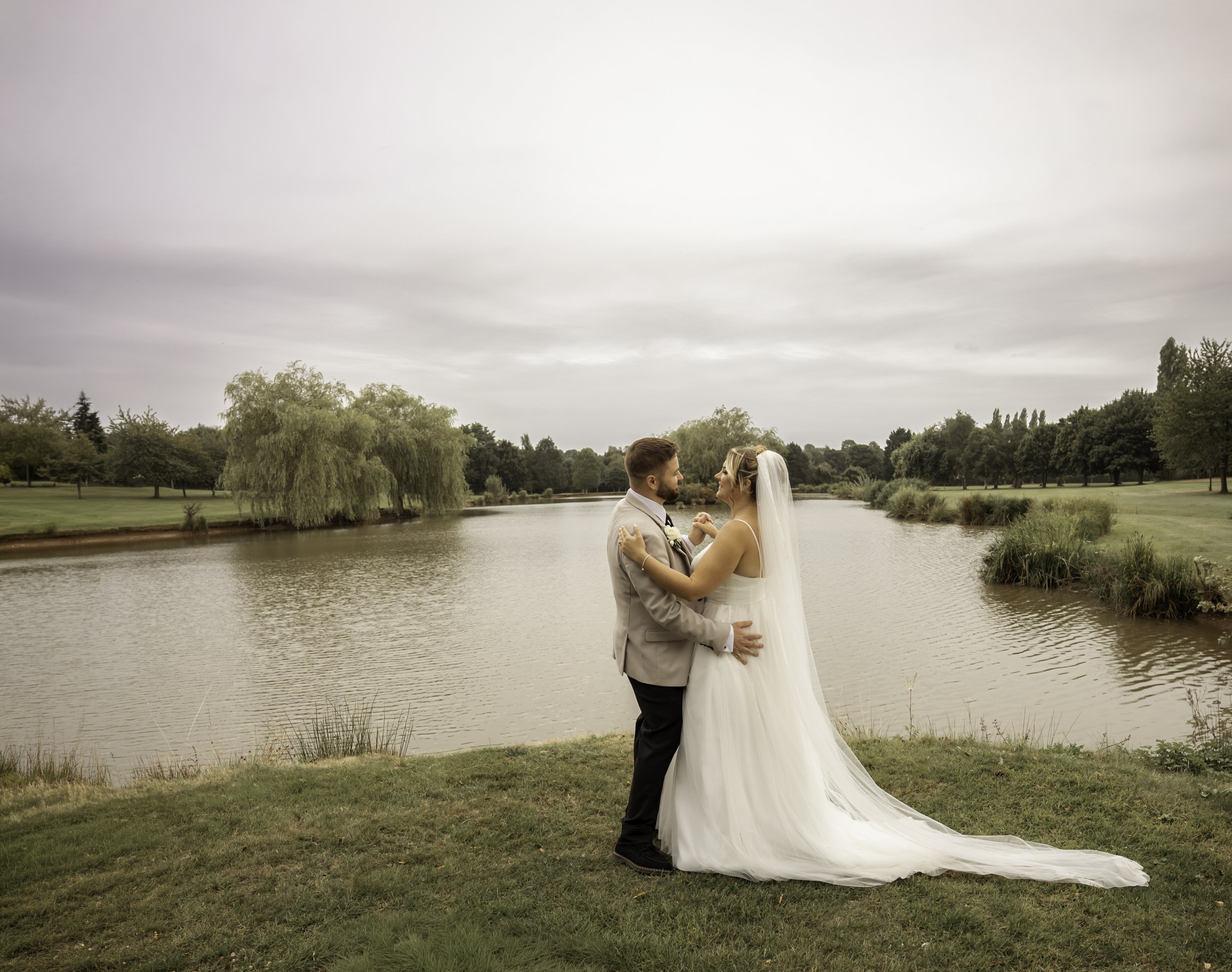 A bride and groom standing by a lake or river, holding hands, surrounded by trees, on a cloudy day.