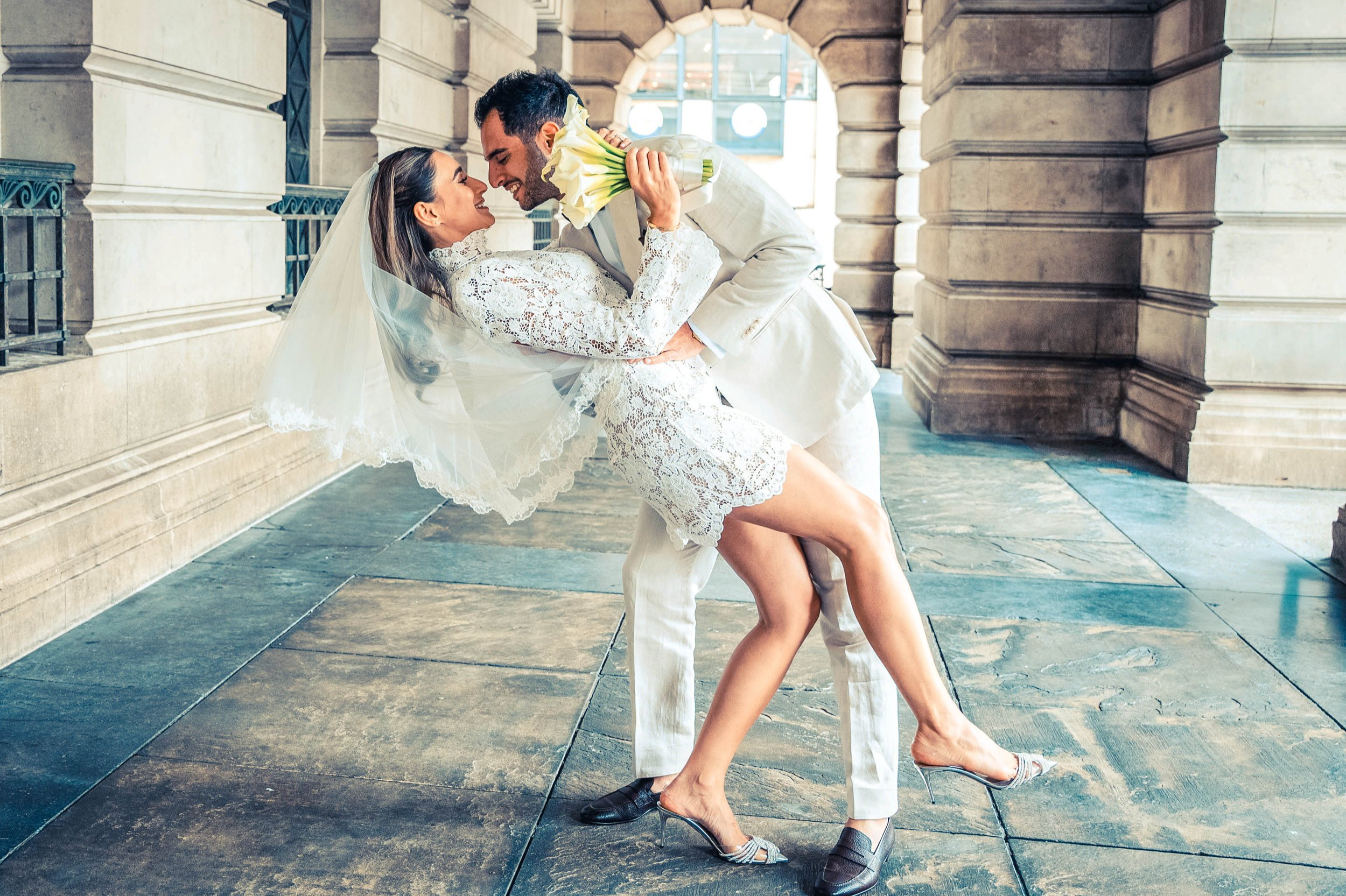 A couple in wedding attire dancing inside a building with stone walls and large windows, smiling and looking at each other, with the man lifting the woman.
