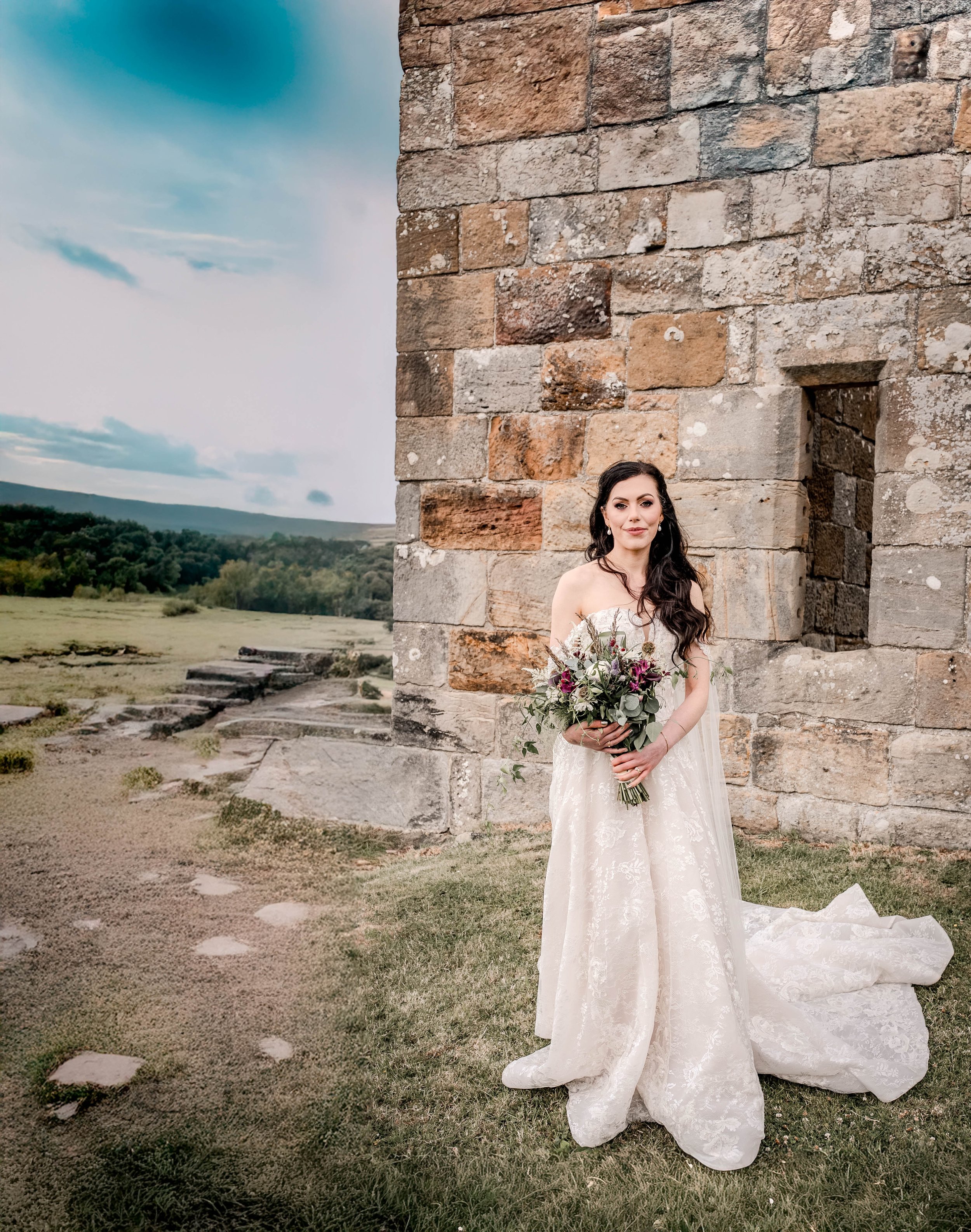 A bride in a white wedding gown holding a bouquet of flowers standing outside next to a stone building, with a scenic landscape and cloudy sky in the background.