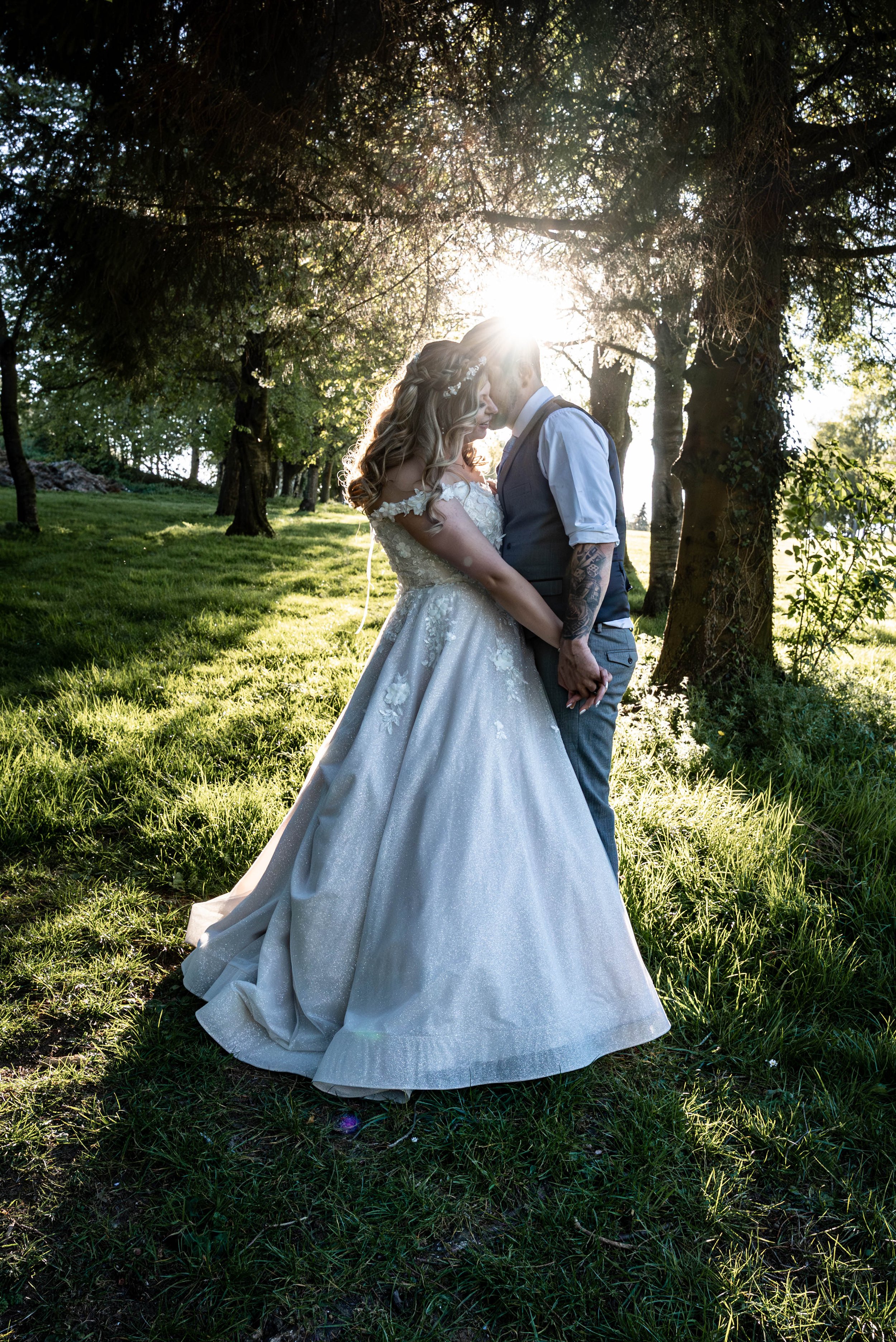 A bride and groom standing close together outdoors, holding hands, with their foreheads touching, illuminated by sunlight filtering through trees.