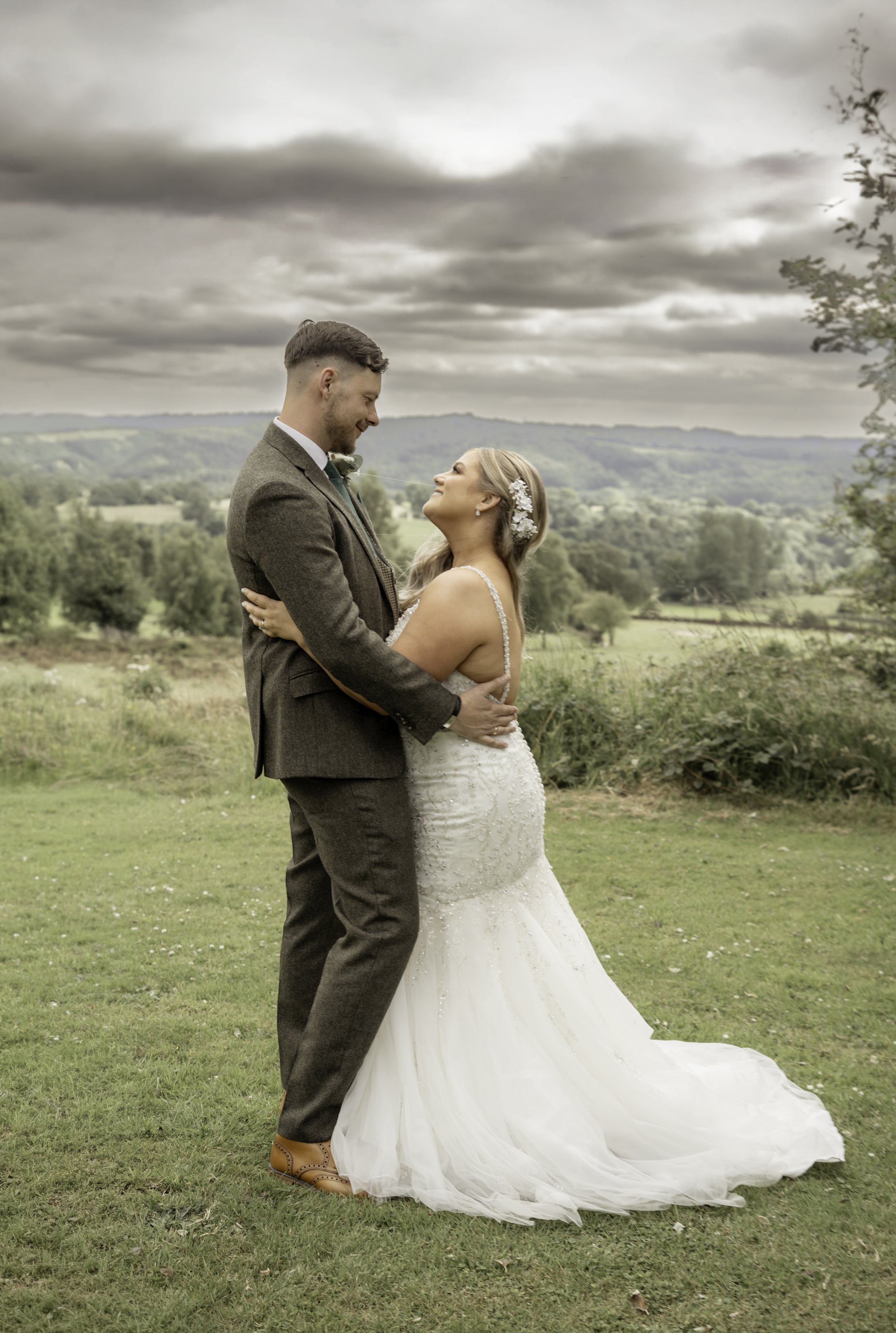 A bride and groom embracing outdoors during their wedding, with a scenic countryside in the background.