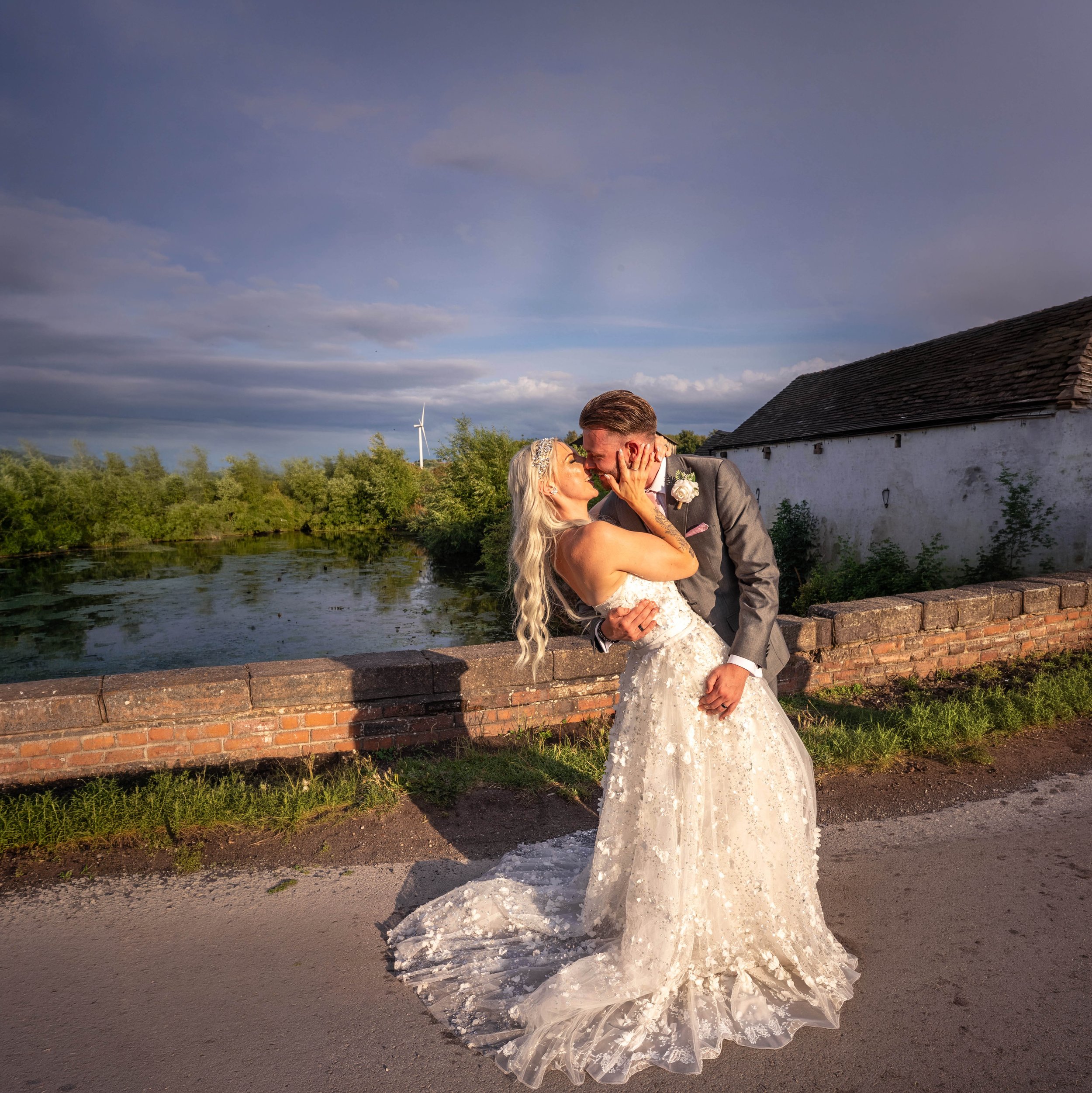A bride and groom share a kiss outdoors near a body of water, with a cloudy sky, a wind turbine, and a white building with a dark roof in the background.