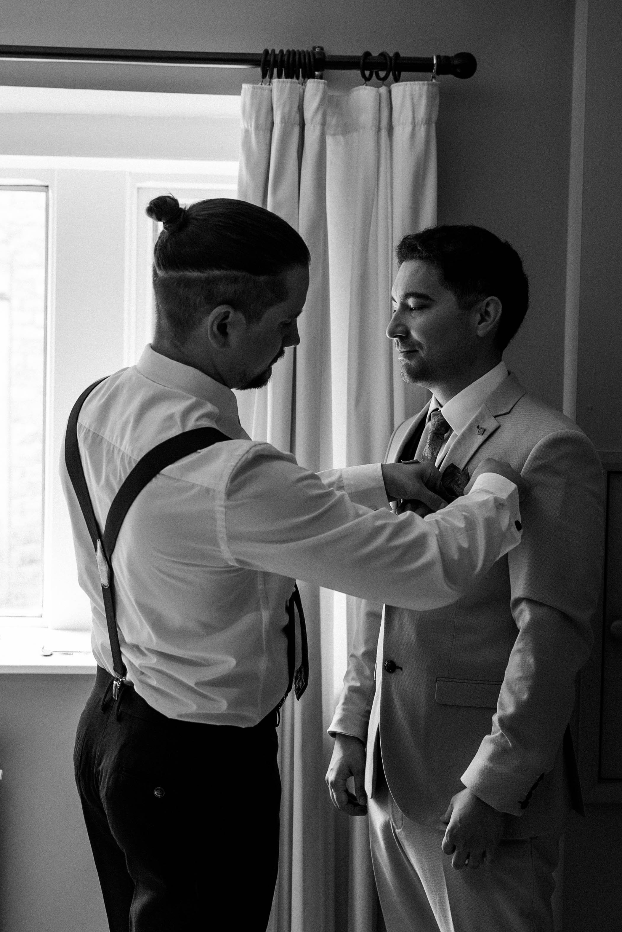 A man in a light-colored suit is being helped with his tie by another man in a white shirt with suspenders, in a room with curtains and a window.