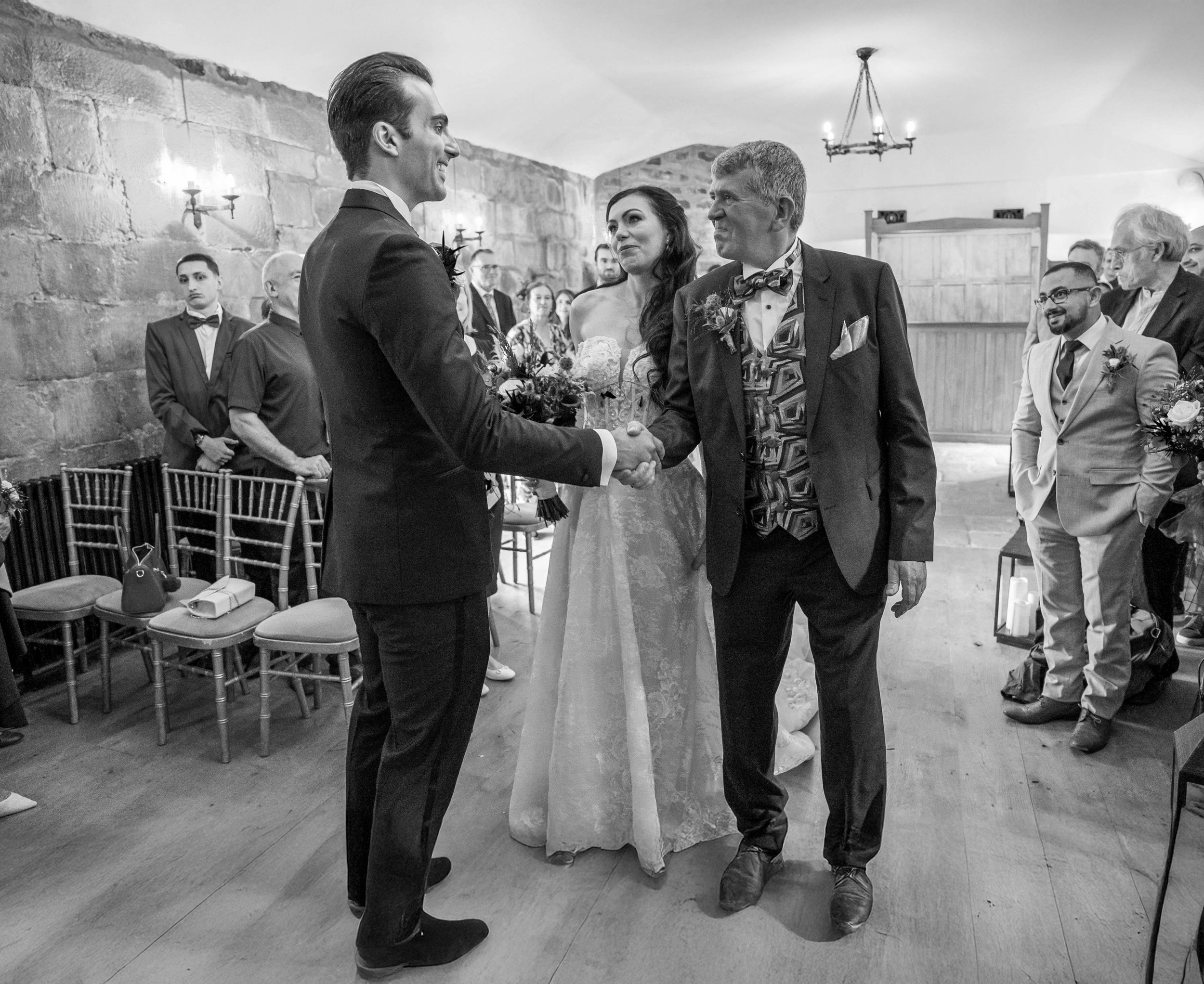 A black and white photo of a wedding ceremony where a groom is shaking hands with an older man. The bride stands next to the groom, holding a bouquet and looking at him. Several guests are seated and standing in the background, watching the event. Th