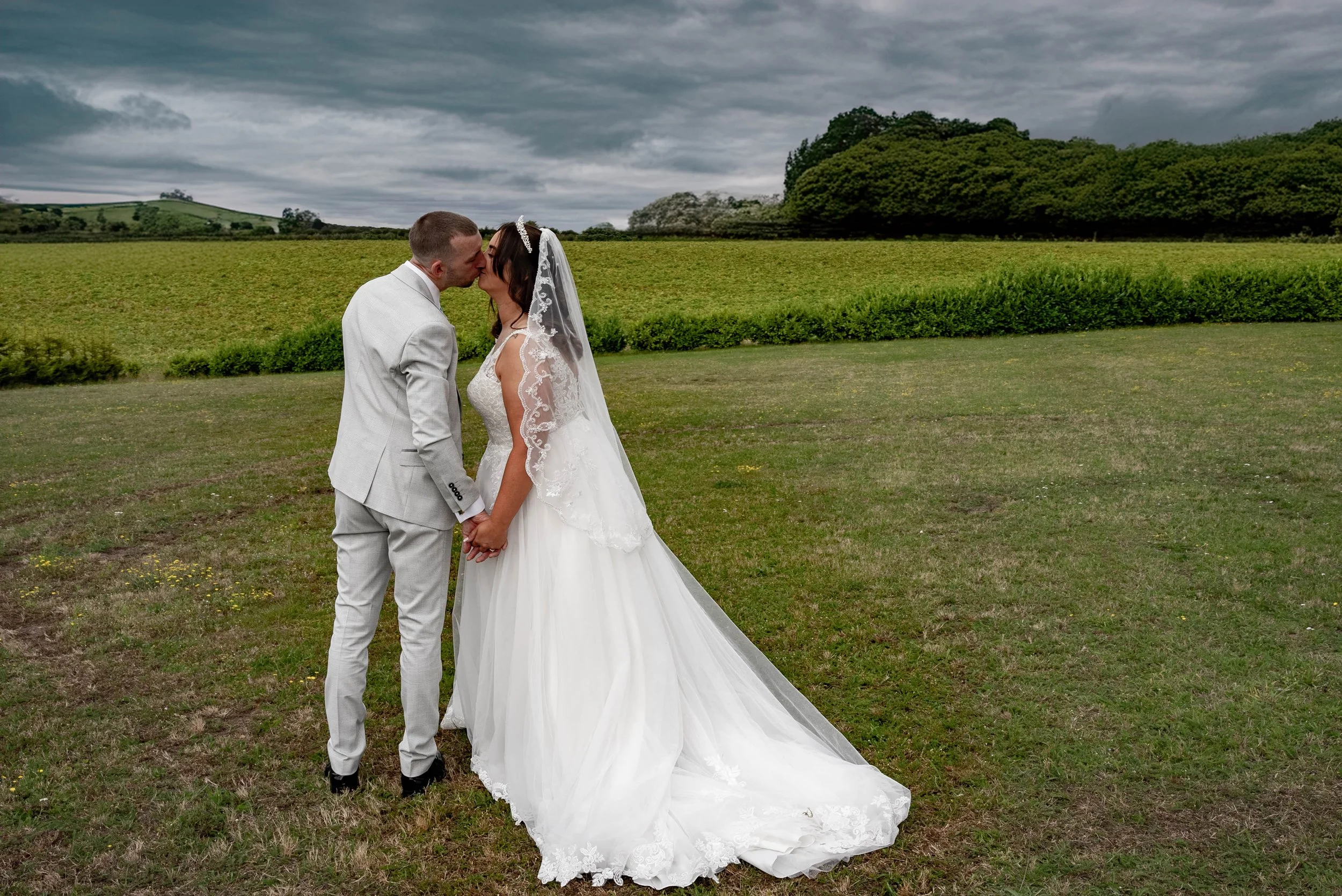 A bride and groom kissing outdoors in an open grassy field with green trees and dark cloudy sky in the background.