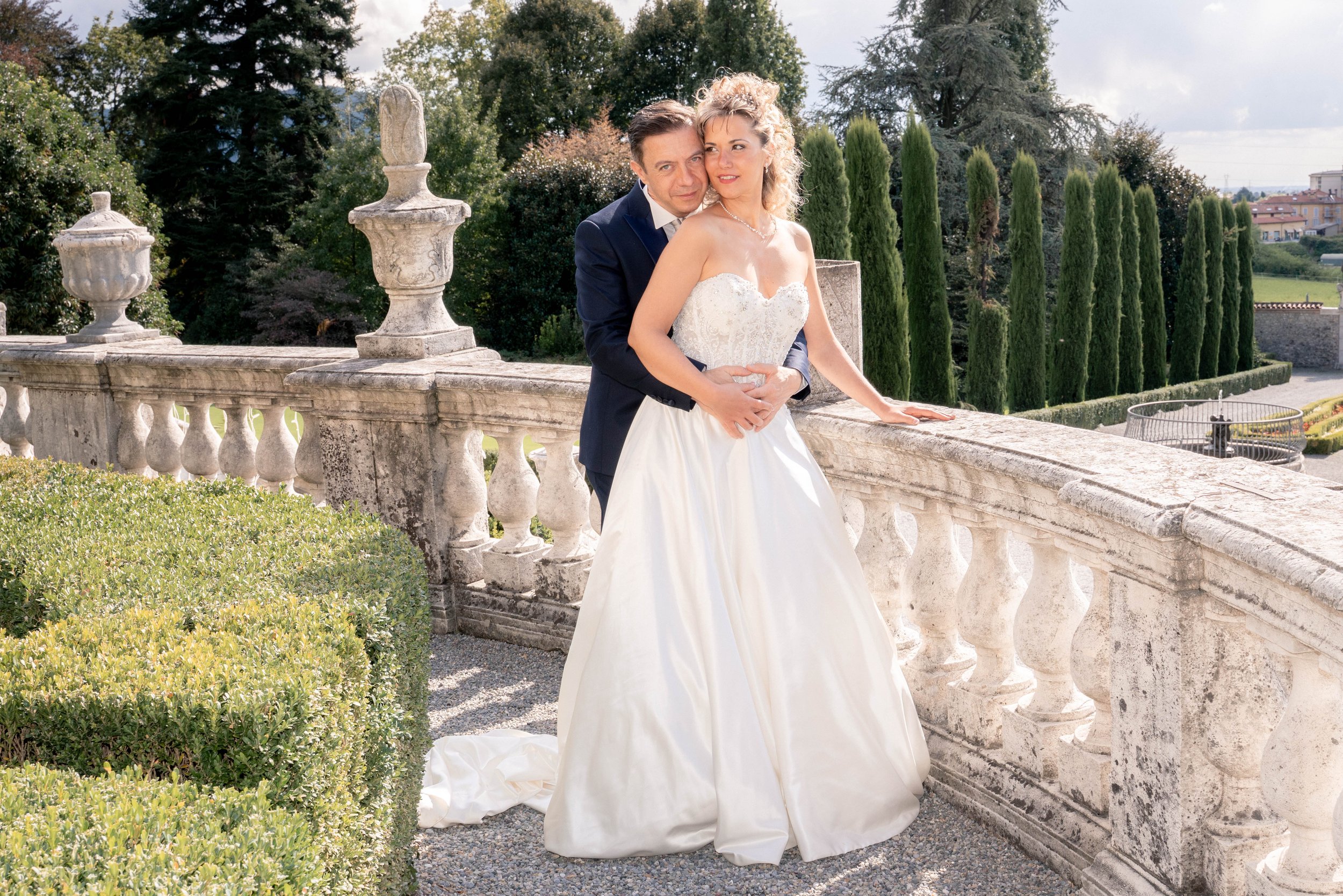 A man and woman in wedding attire standing on a stone balcony with greenery and trees in the background.