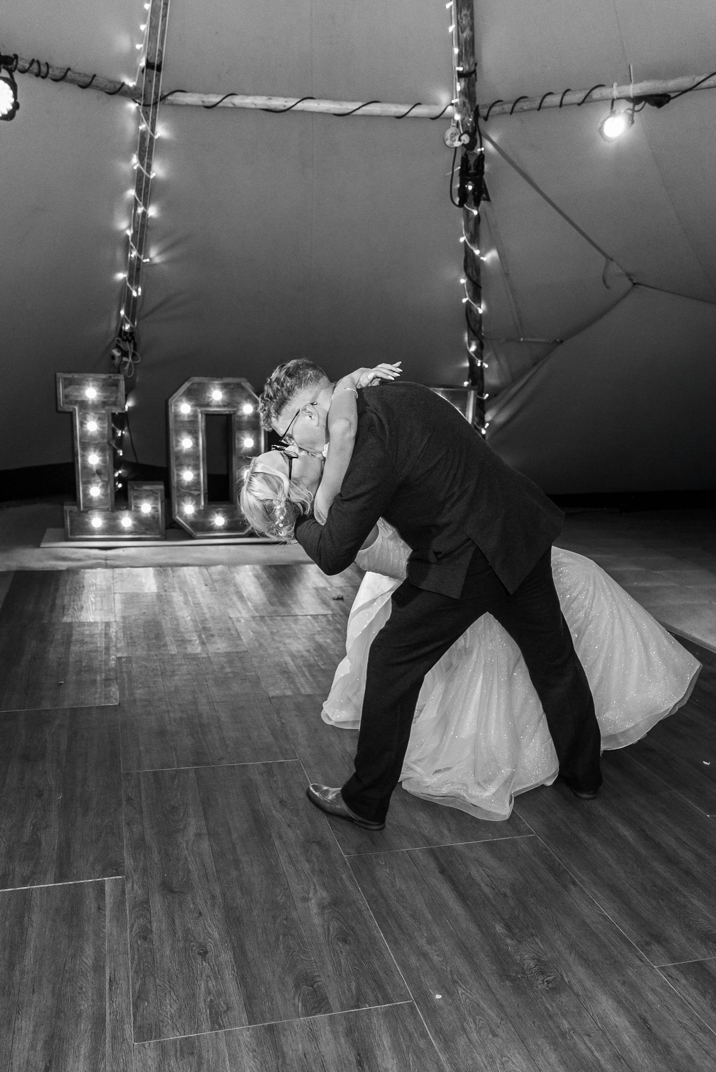 A couple sharing a dance at a wedding reception, with large illuminated letters "LOVE" in the background and string lights hanging from the ceiling.