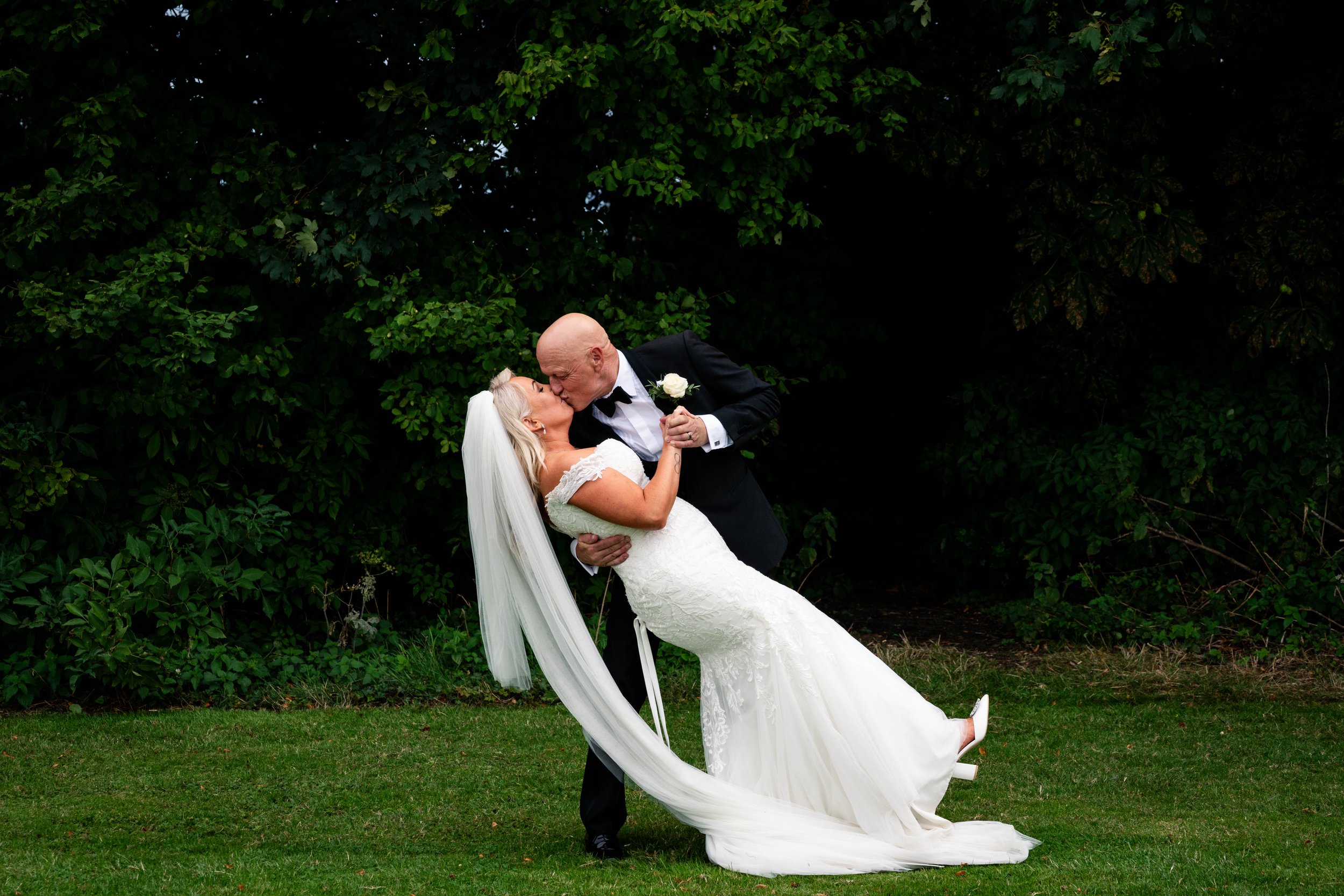 A groom lifting a bride in a wedding dress as they kiss outdoors with green trees in the background.