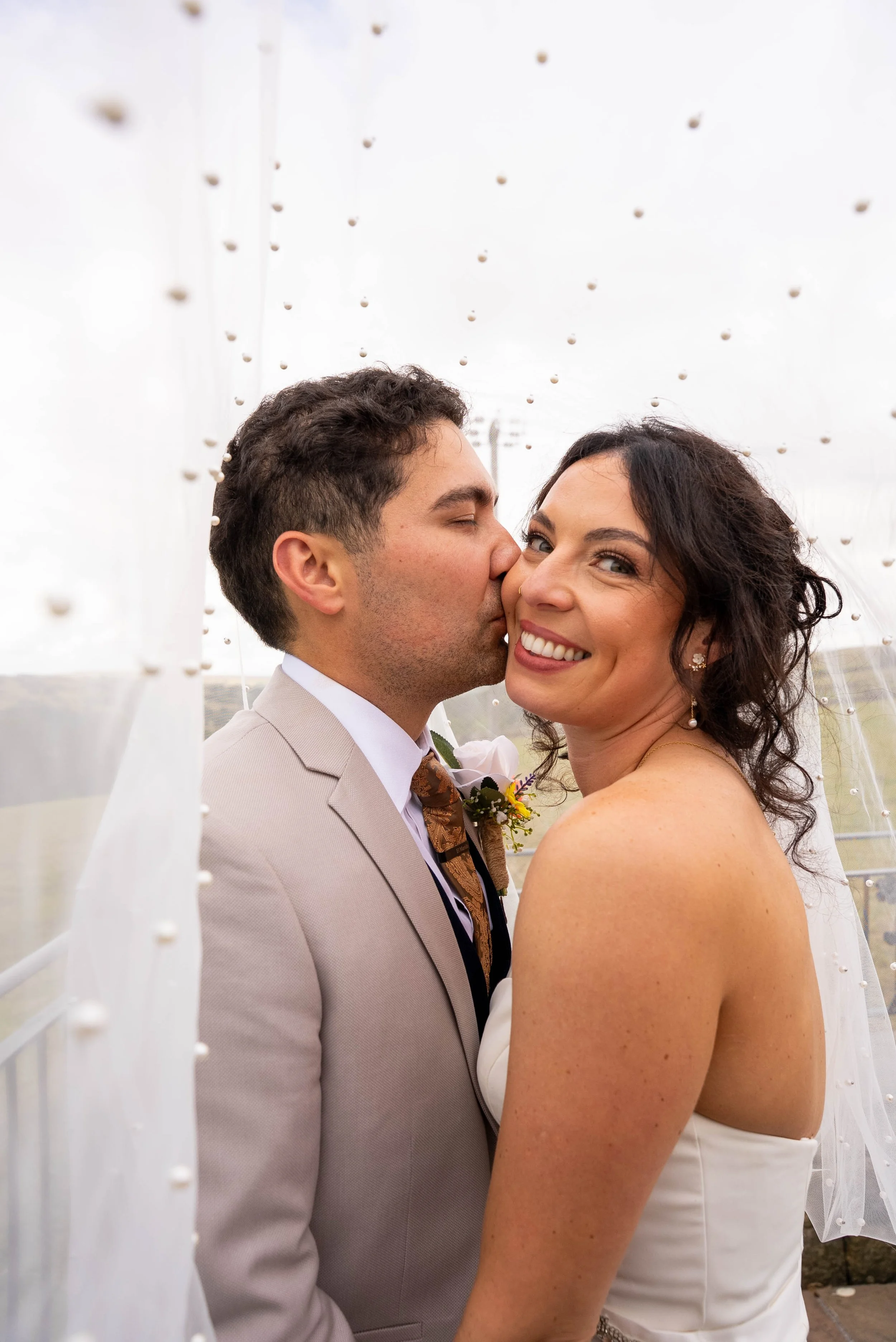 A newlywed couple shares a kiss outdoors during their wedding, with the groom kissing the bride on the cheek. The bride is smiling and looking at the camera, wearing earrings and a strapless white wedding dress, while the groom is in a light-colored 