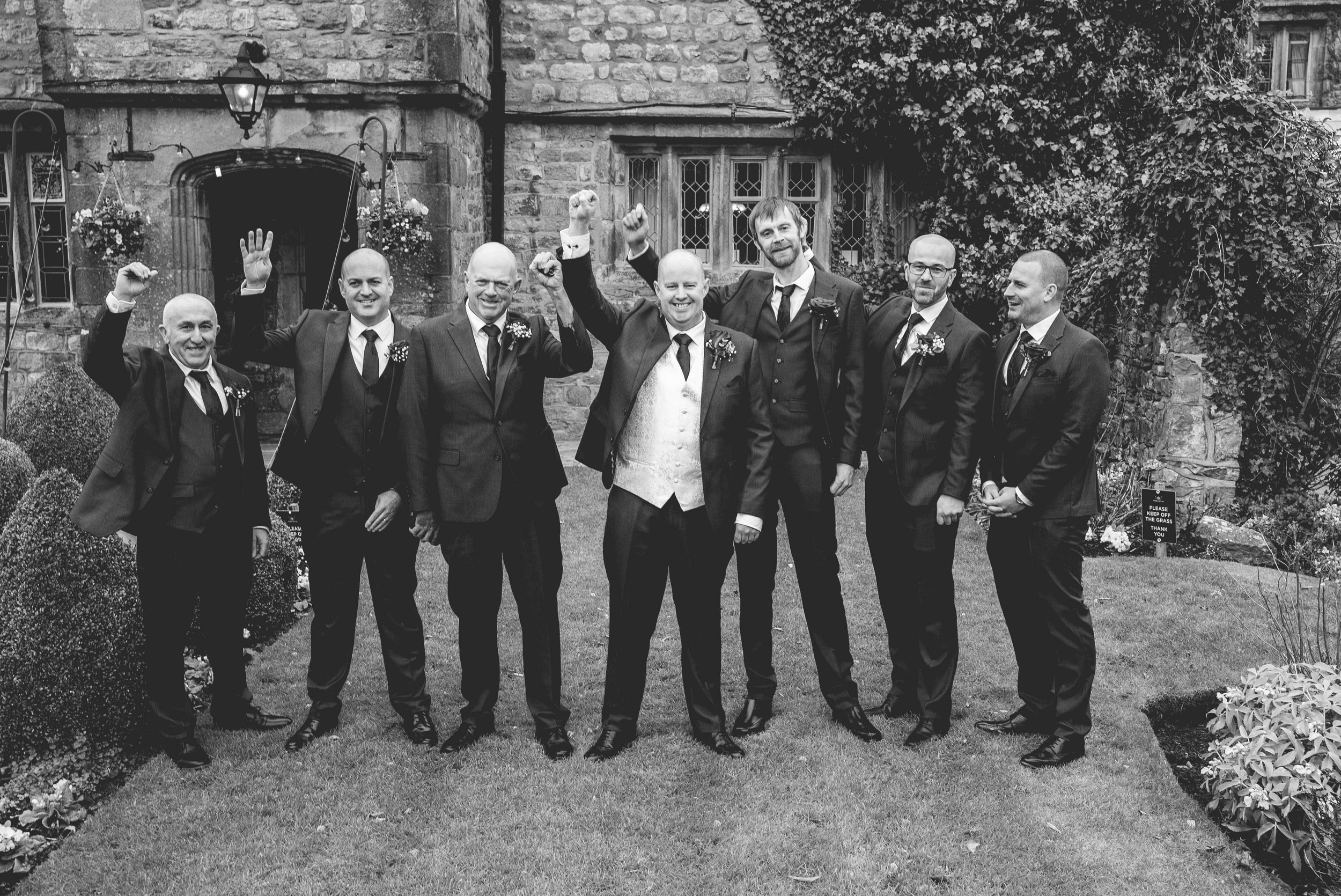 Group of seven men dressed in suits posing outside in front of a stone building, celebrating with raised fists.