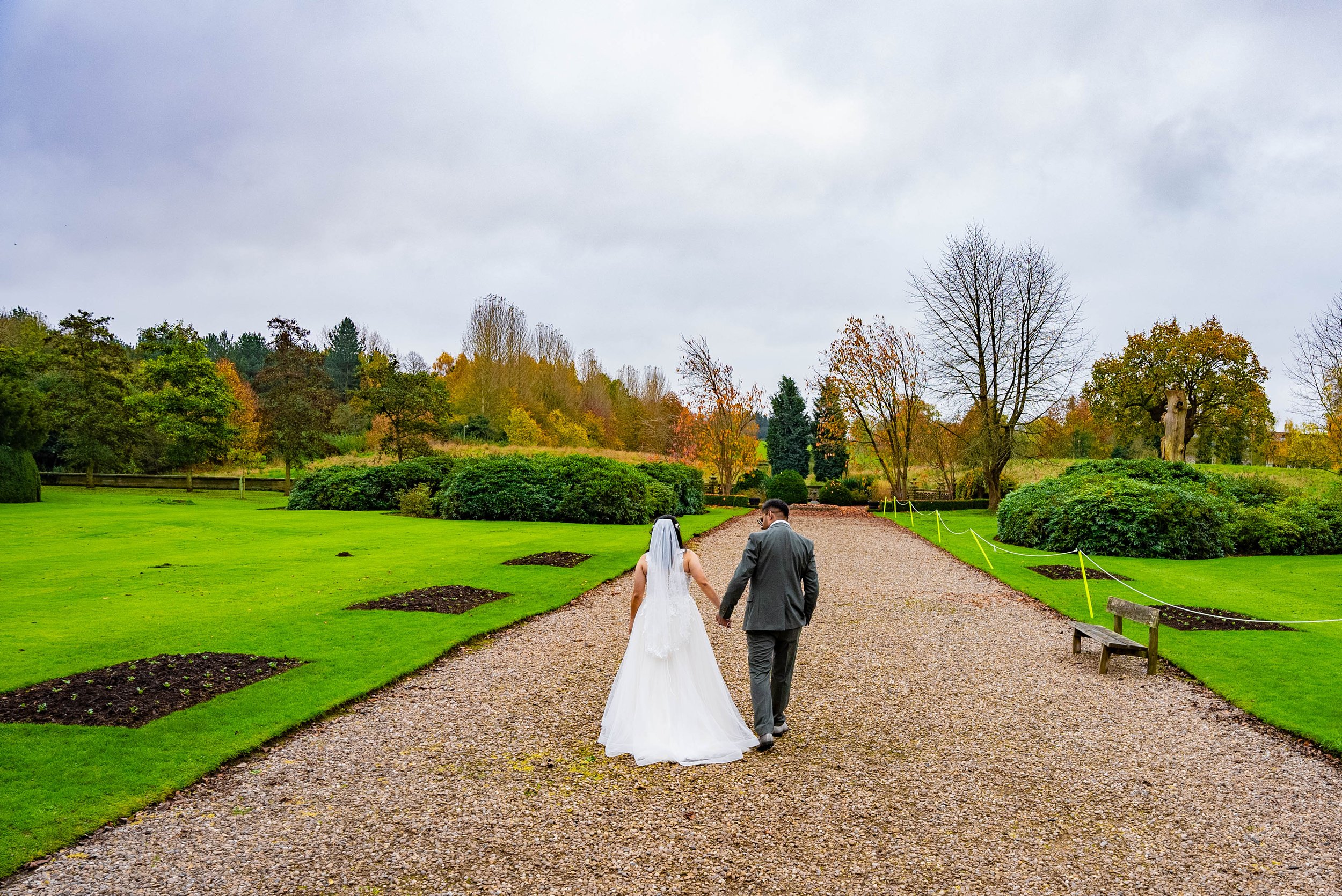 A bride and groom walking hand in hand on a gravel pathway in a park during autumn, with green grass, bushes, and trees in the background, on a cloudy day.