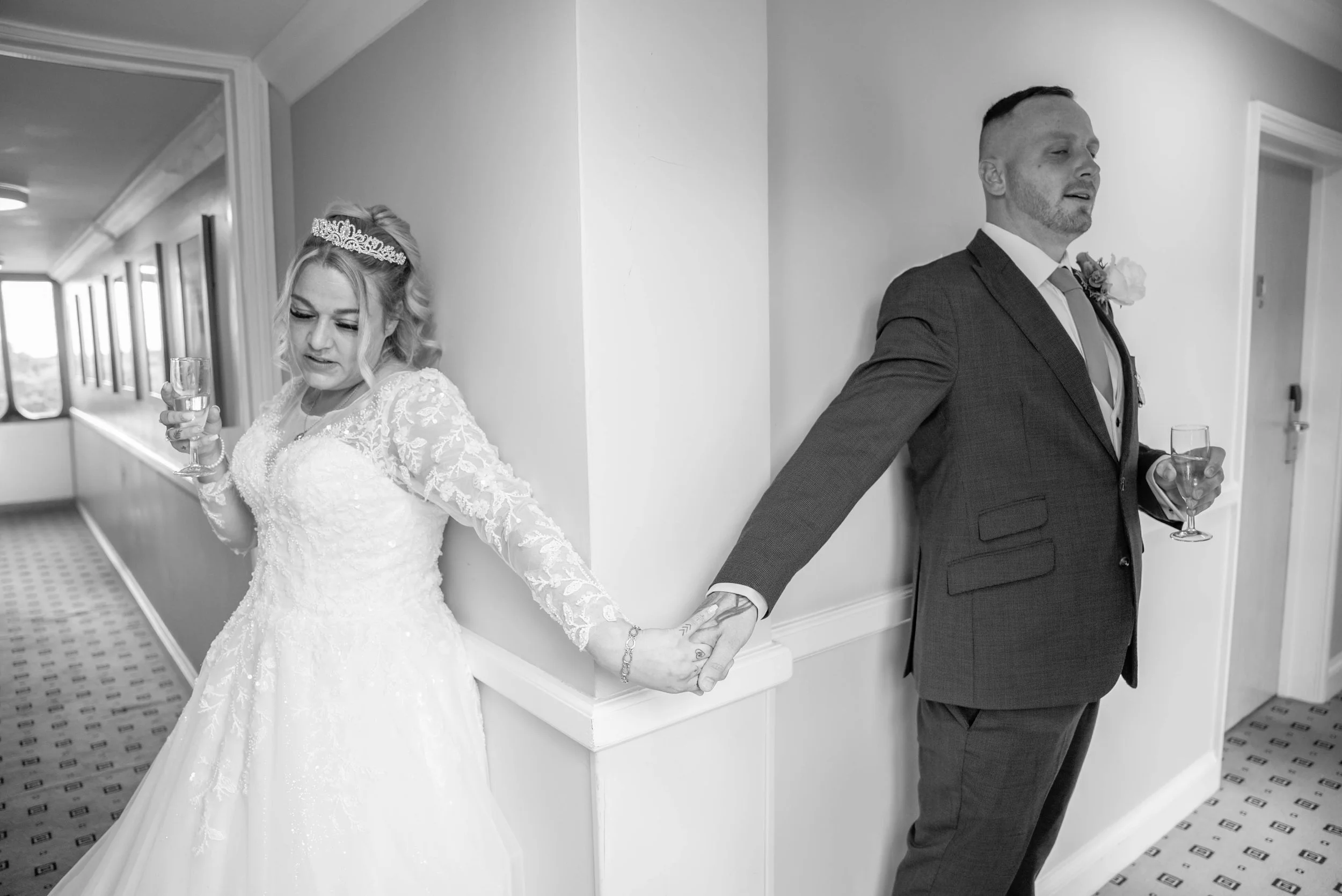 A bride and groom stand back-to-back, holding hands behind their backs, each holding a glass of champagne, in a corridor during their wedding celebration.