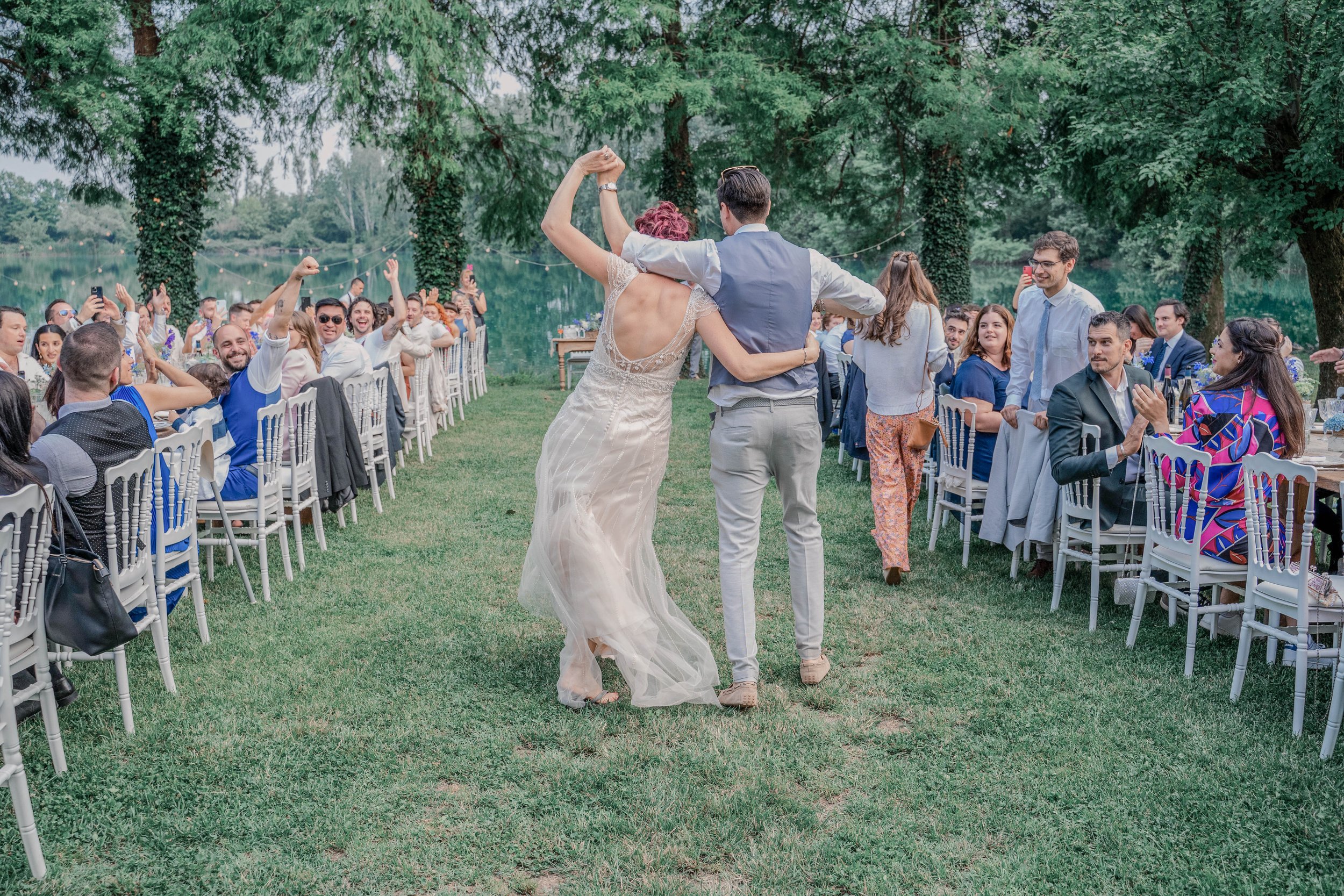 Bride and groom dancing together at an outdoor wedding reception by a lake, surrounded by seated guests, under tall green trees.