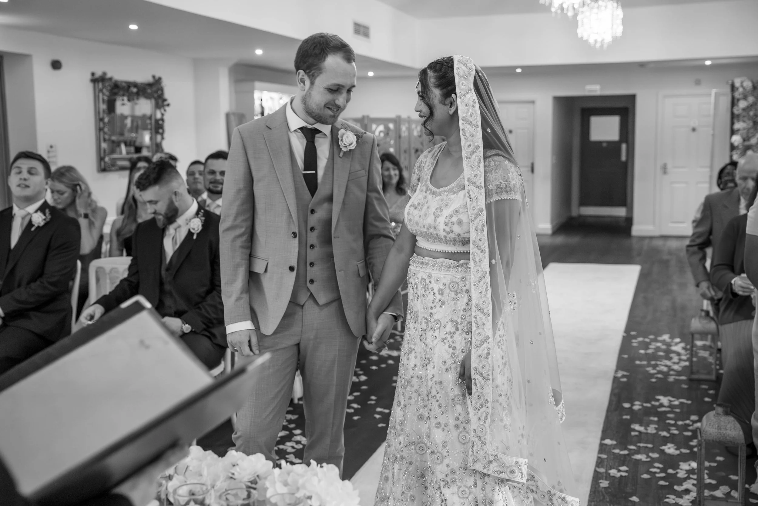 A bride and groom holding hands during their wedding ceremony inside a decorated room, surrounded by seated guests.