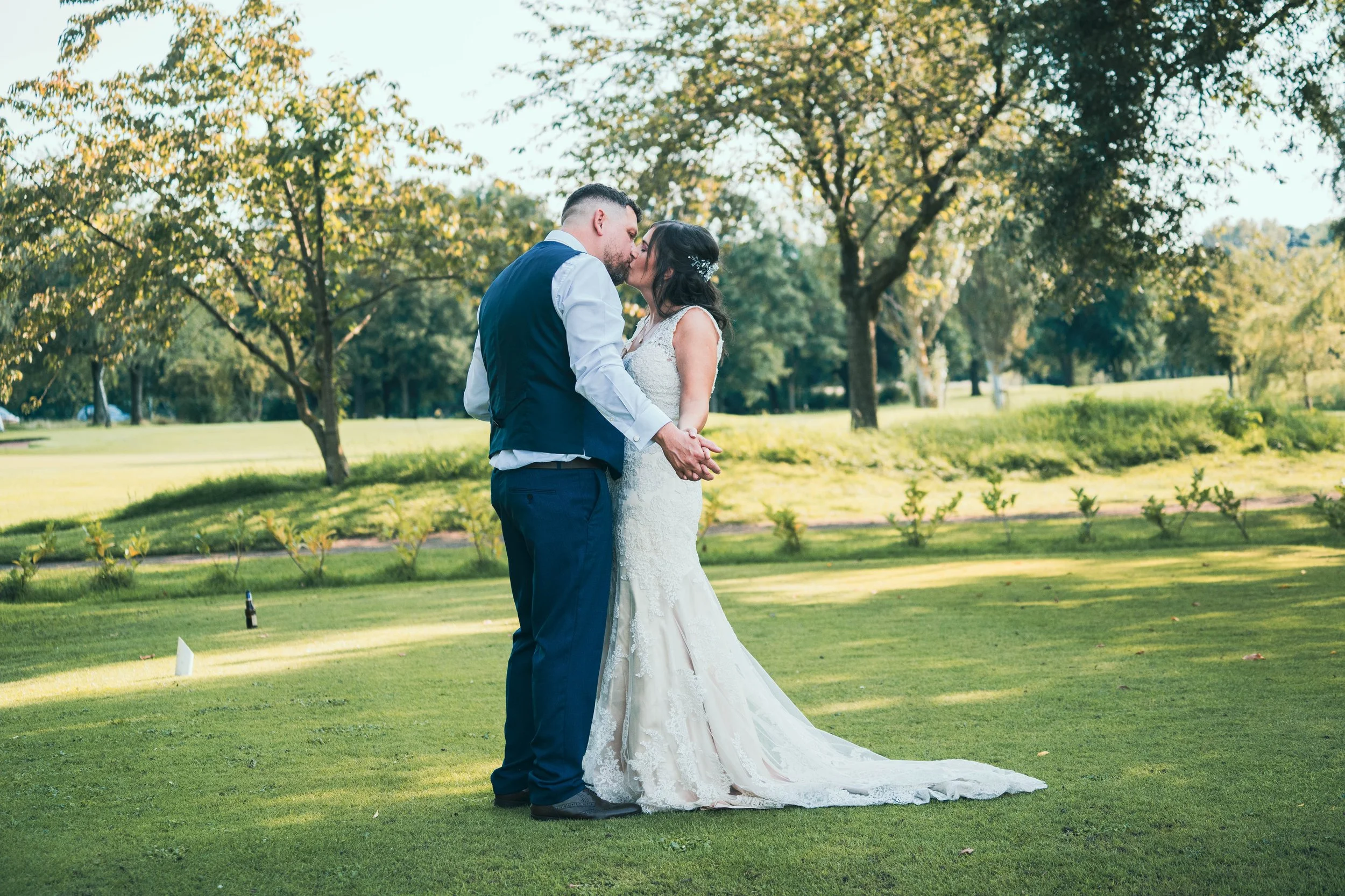 A bride and groom kissing outdoors on a grassy field with trees in the background during their wedding.