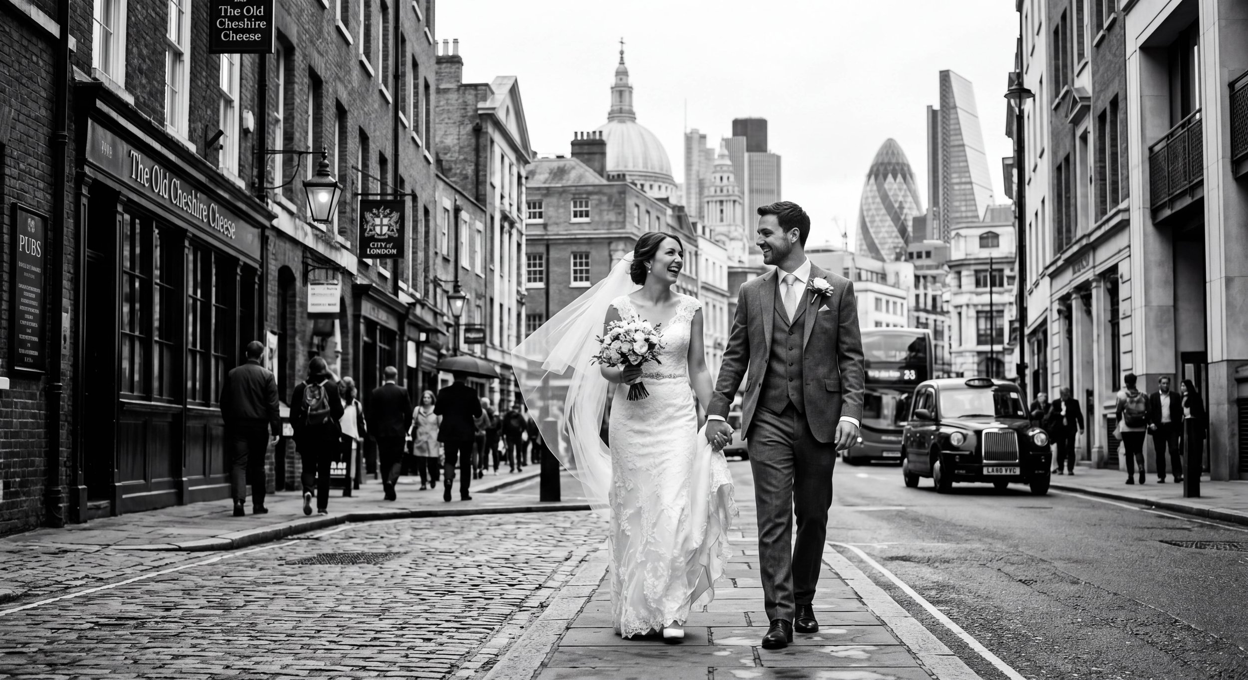 A black-and-white photo of a smiling bride and groom walking hand-in-hand on a city street. The bride is wearing a wedding dress with lace details and holding a bouquet. The groom is in a suit with a boutonniere. The street has pedestrians, cars, and