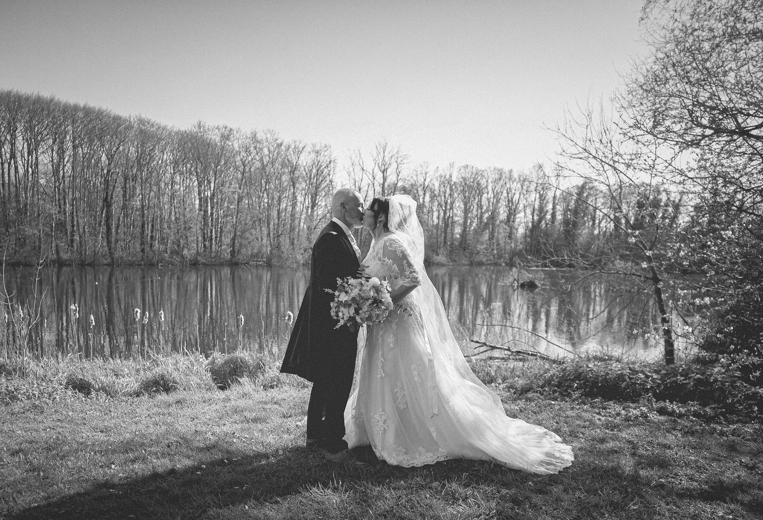 A black-and-white photo of a bride and groom kissing outdoors near a lake, with leafless trees in the background.