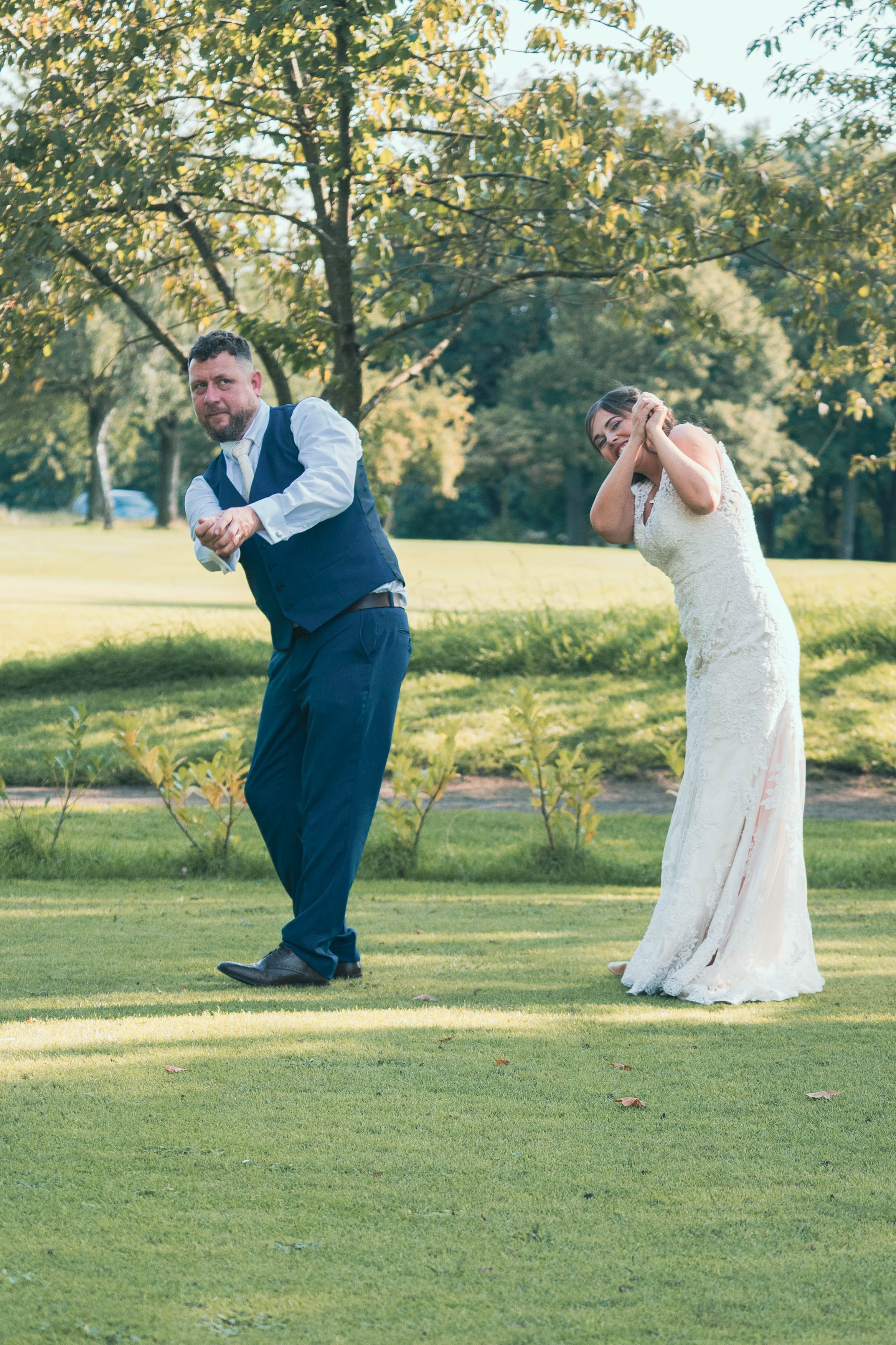 A man and woman in wedding attire outdoors on a sunny day, playfully pointing and hiding behind each other.