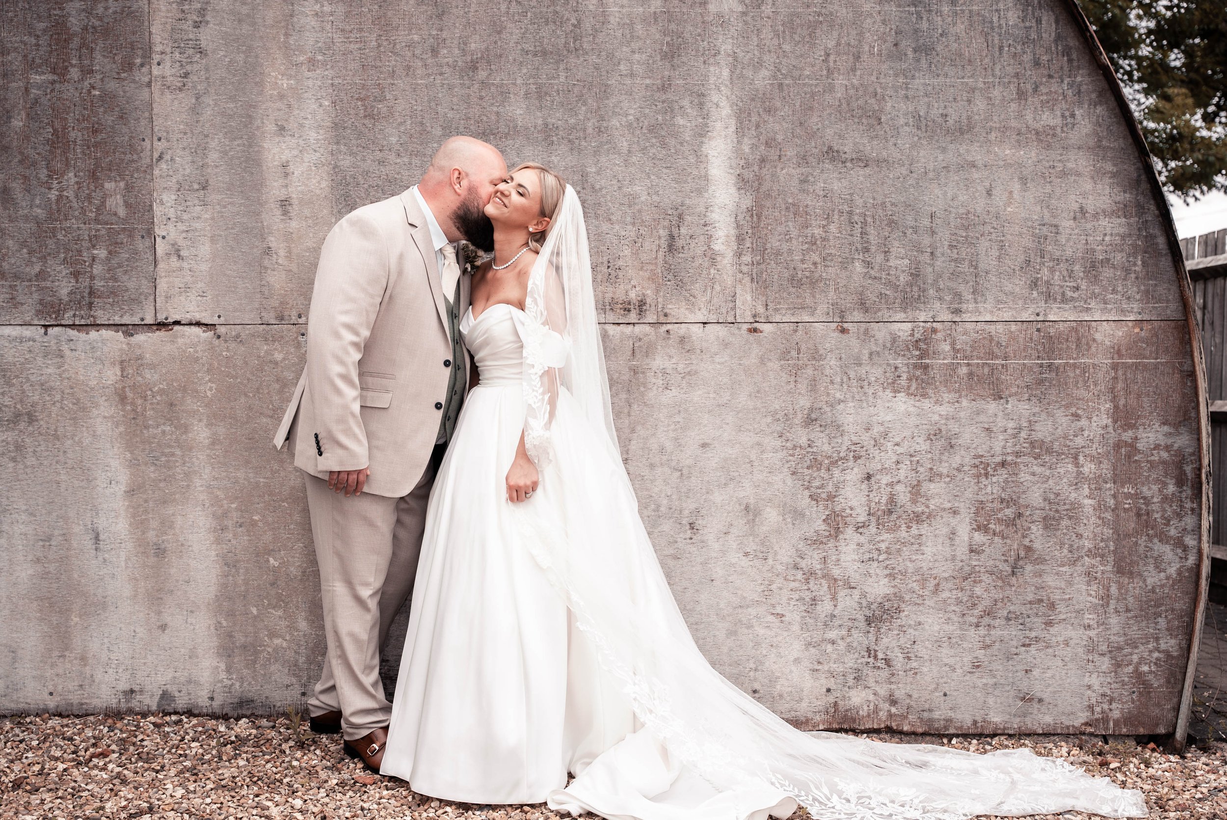 A couple, dressed in wedding attire, sharing a kiss and smiling against a large curved concrete wall.