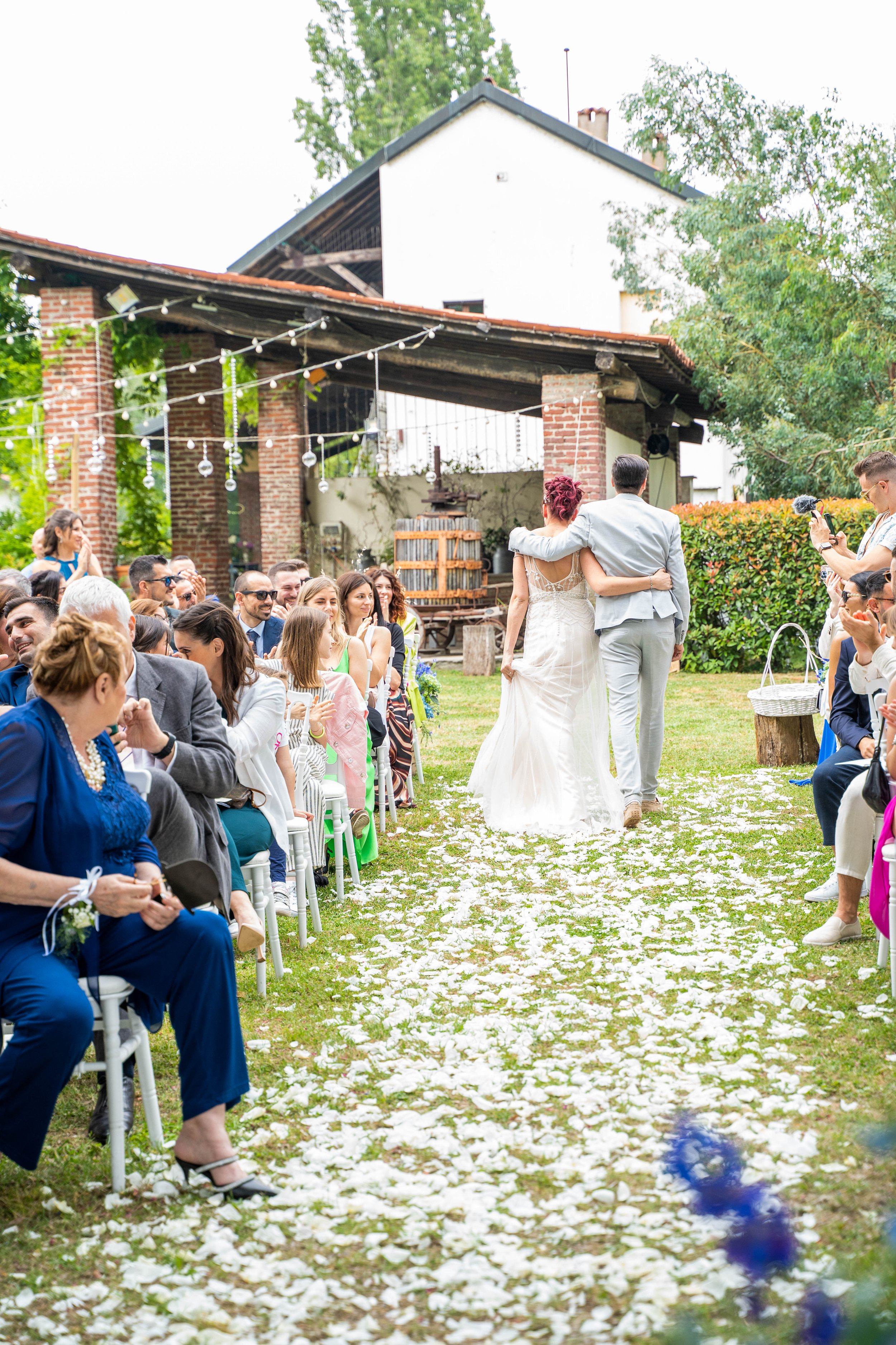 Couple walking down the aisle at an outdoor wedding ceremony, surrounded by seated guests, decorated with flower petals on the ground and string lights overhead.
