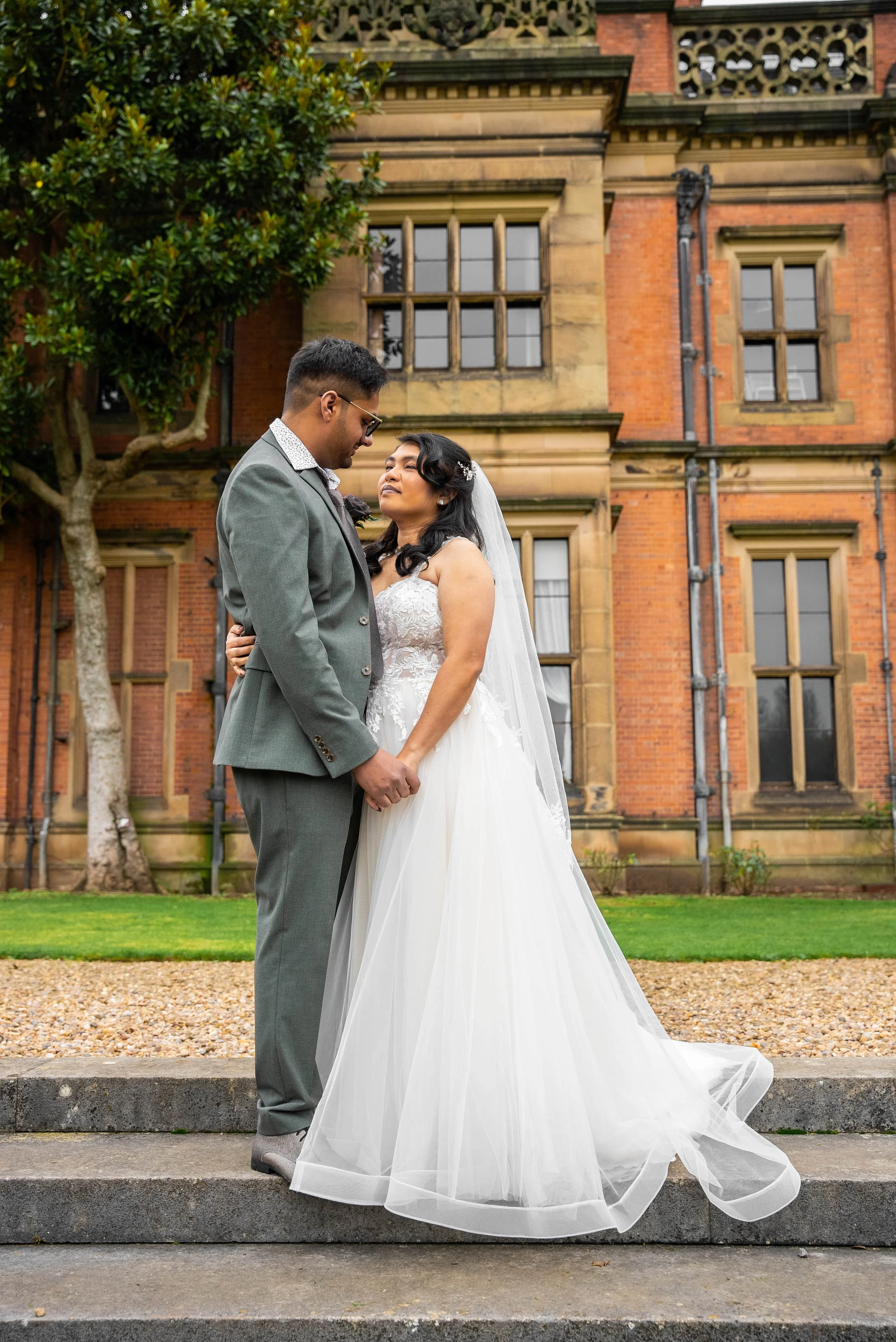 A bride and groom standing on steps outside a brick building, holding hands and looking into each other's eyes.