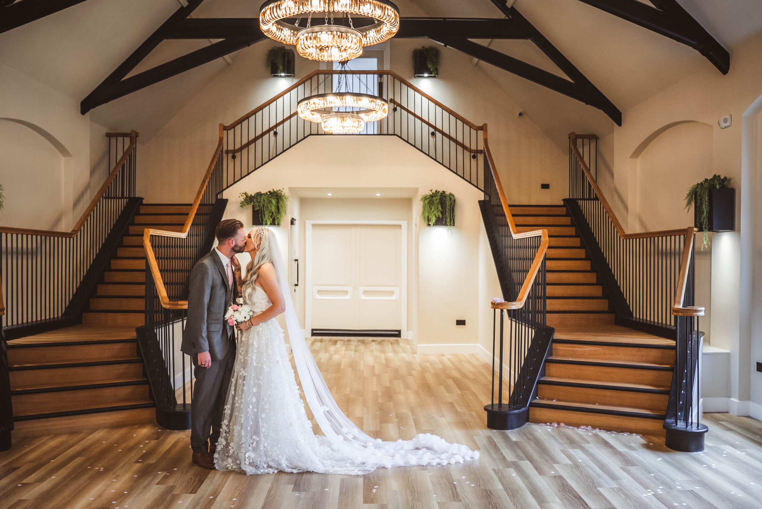 A bride and groom sharing a kiss in an elegant indoor venue with a grand staircase, chandelier, and minimal decor.