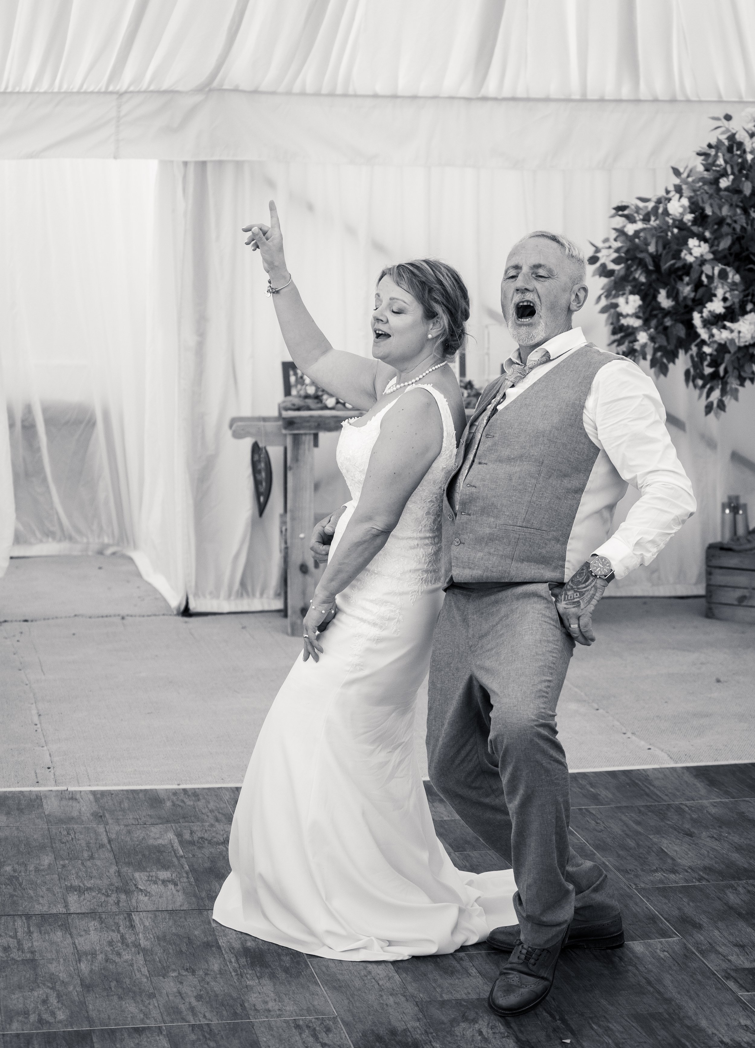 An elderly couple dancing at a wedding reception. The woman is wearing a white wedding dress and pearl necklace, and the man is in a vest and tie. They are smiling and appear to be enjoying themselves.