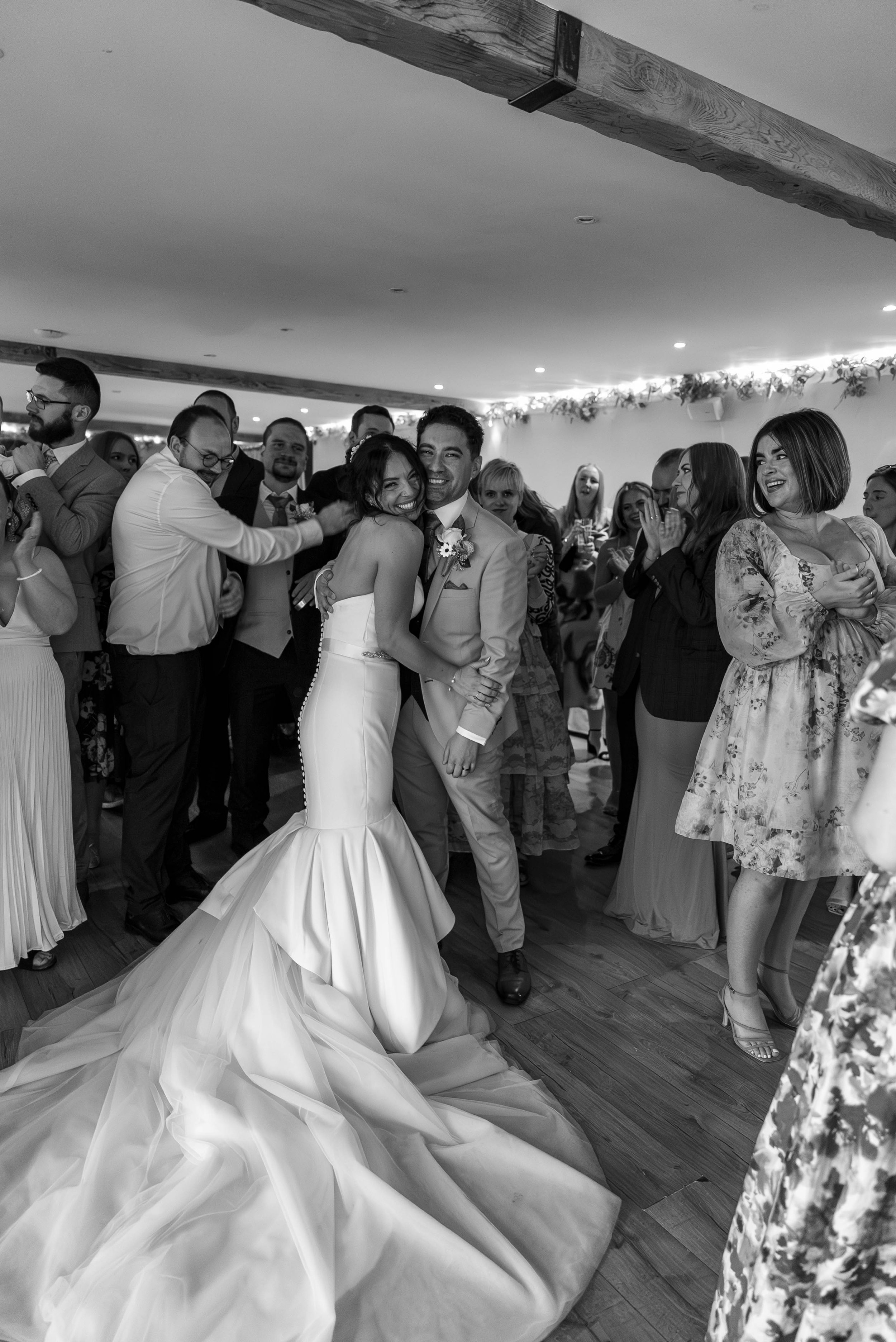 Black and white photo of a wedding reception with a bride and groom in the center, smiling and dancing together, surrounded by guests clapping and celebrating.