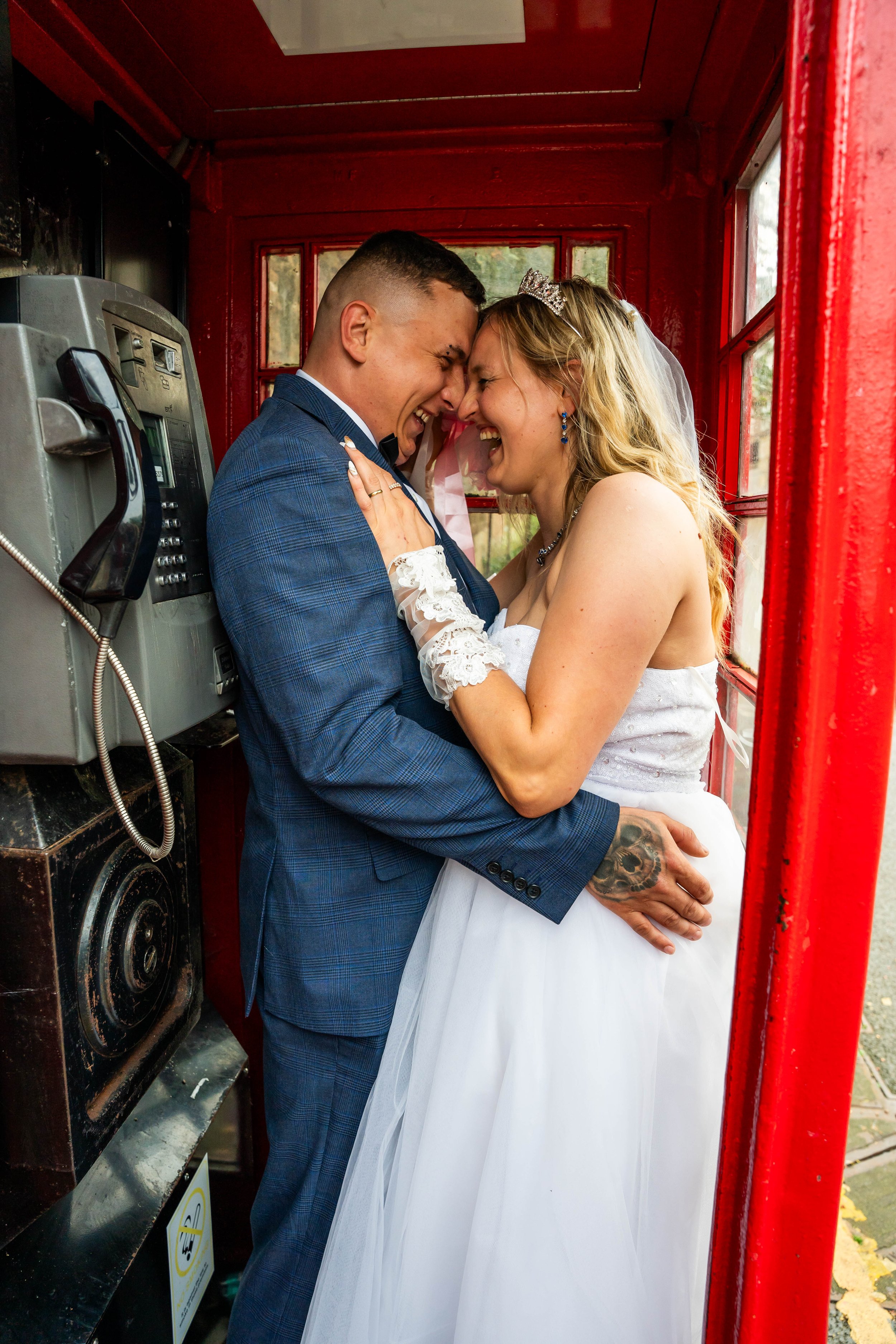 A bride and groom sharing an intimate moment inside a red phone booth, with the groom wearing a blue suit and the bride in a wedding dress, both smiling with their foreheads touching.