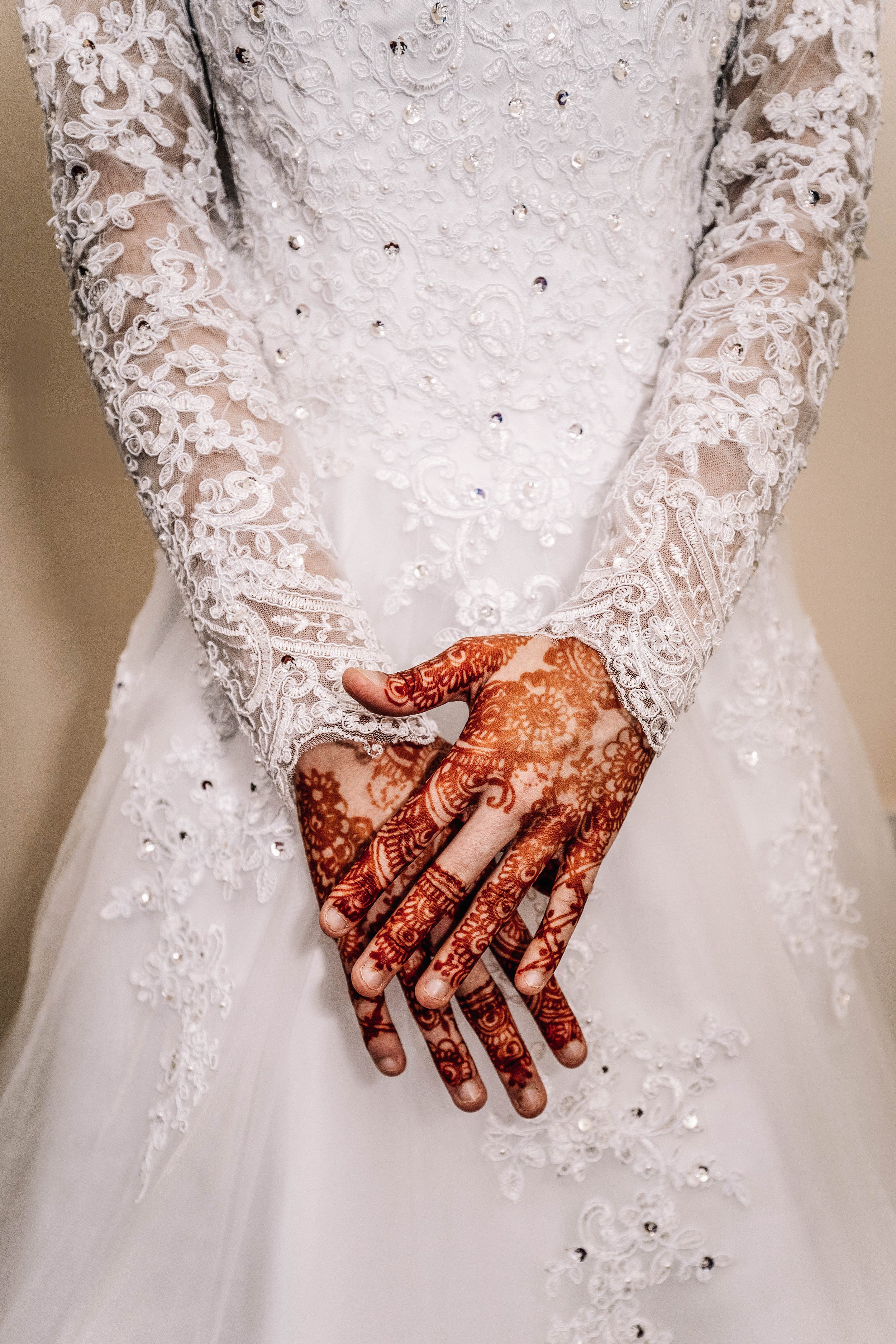 Close-up of a bride's hands with intricate henna designs, wearing a white wedding dress with lace and embroidery details.