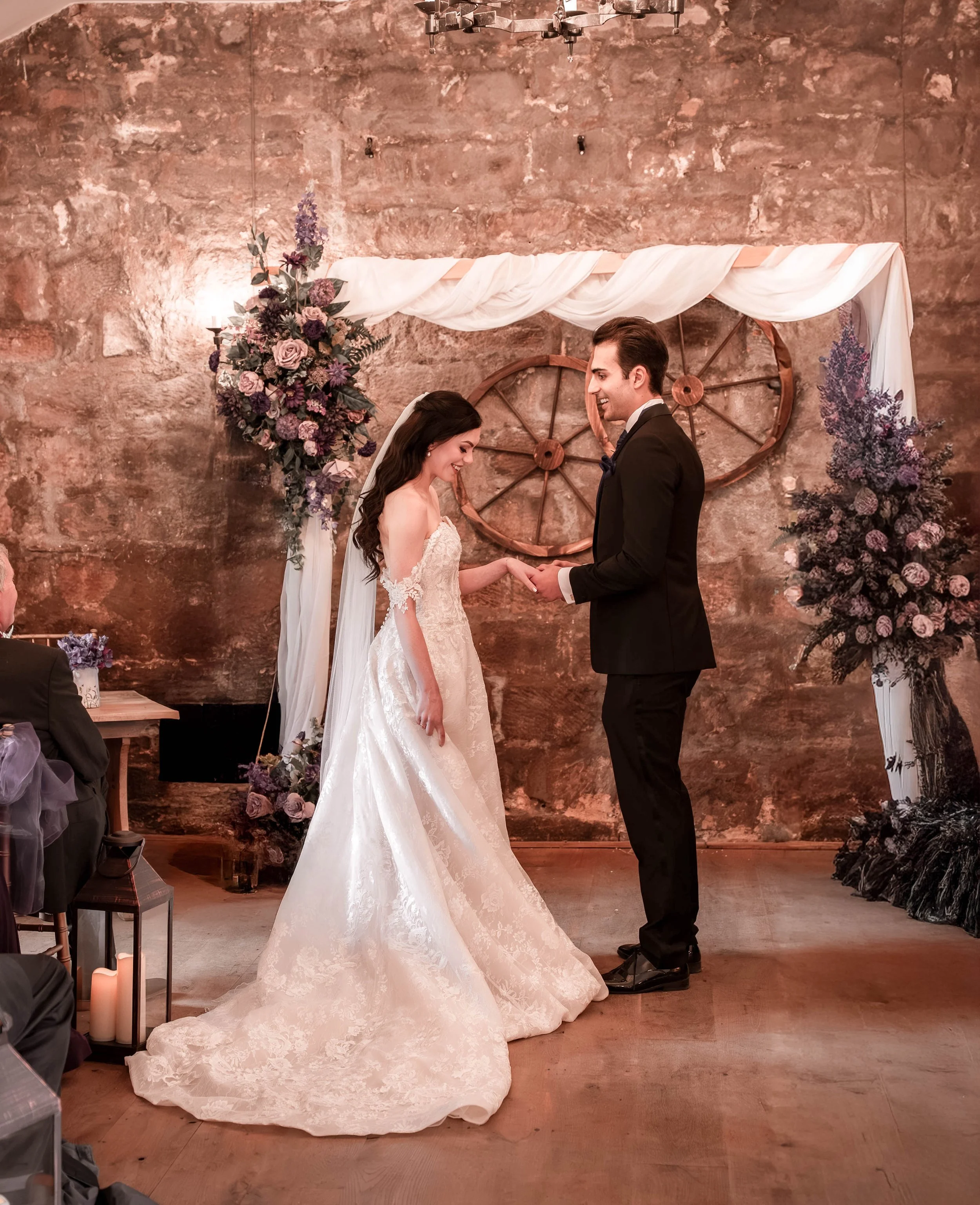 Bride and groom holding hands during their wedding ceremony, standing in front of a rustic backdrop with wheel decorations, floral arrangements, and white drapery.