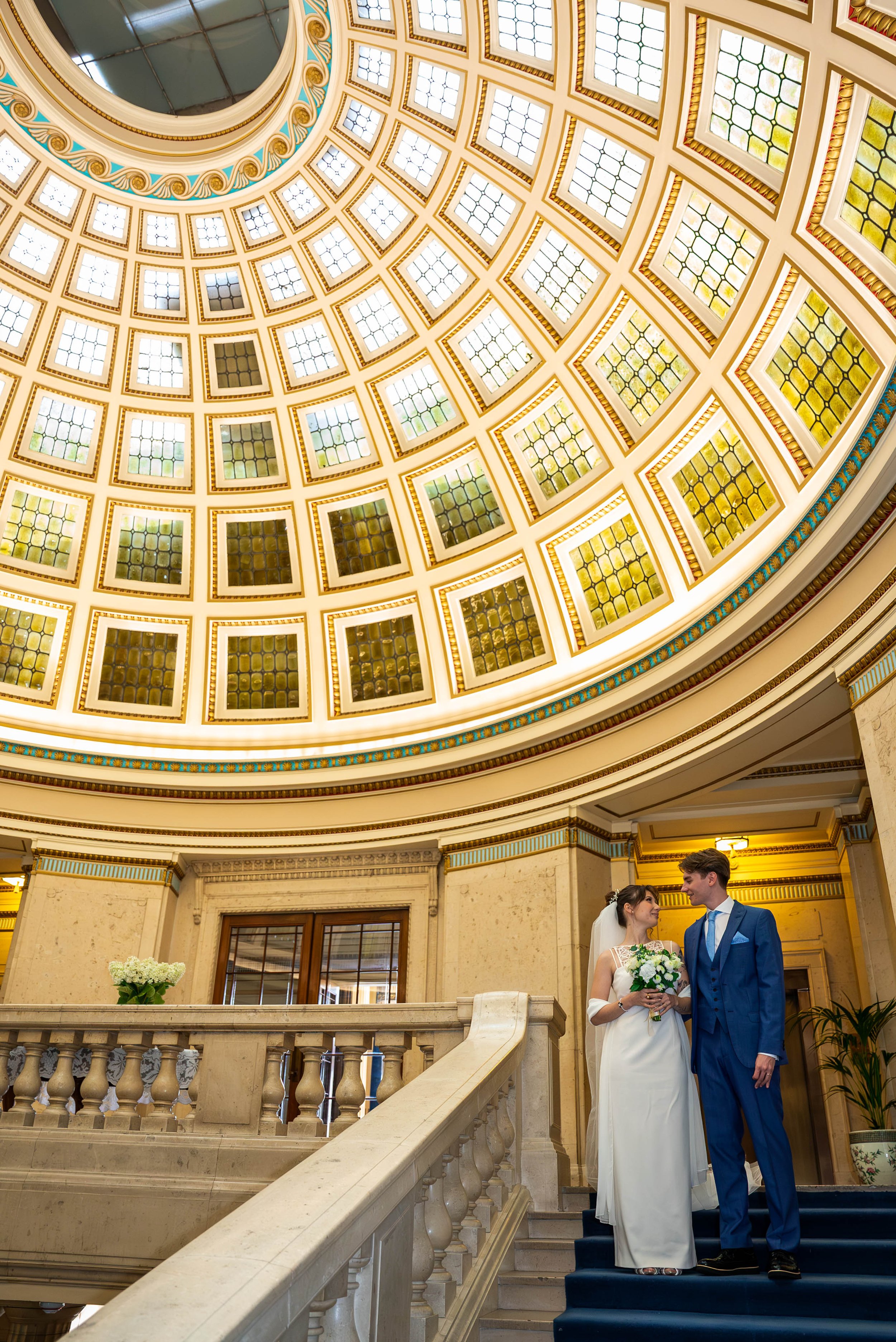 A bride and groom standing on a staircase inside a historic building with a large glass-domed ceiling overhead, decorated with ornate gold and blue accents.