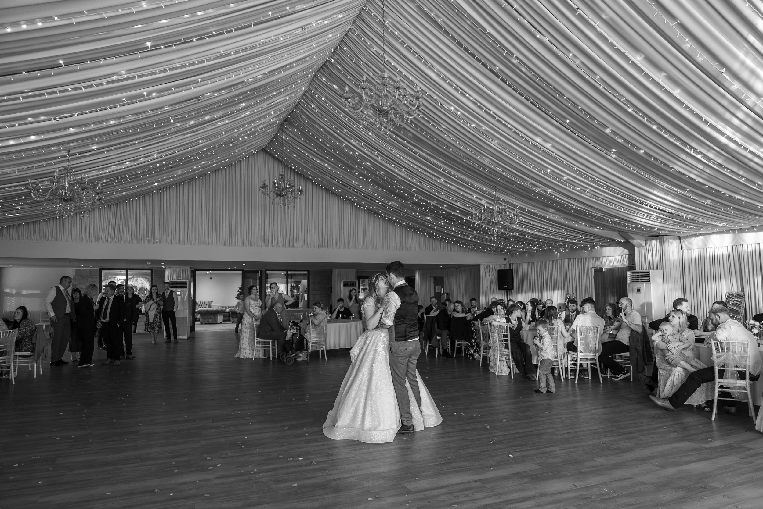 A bride and groom dance in the center of a wedding reception hall with a decorated ceiling and chandeliers, surrounded by guests sitting and standing at tables.