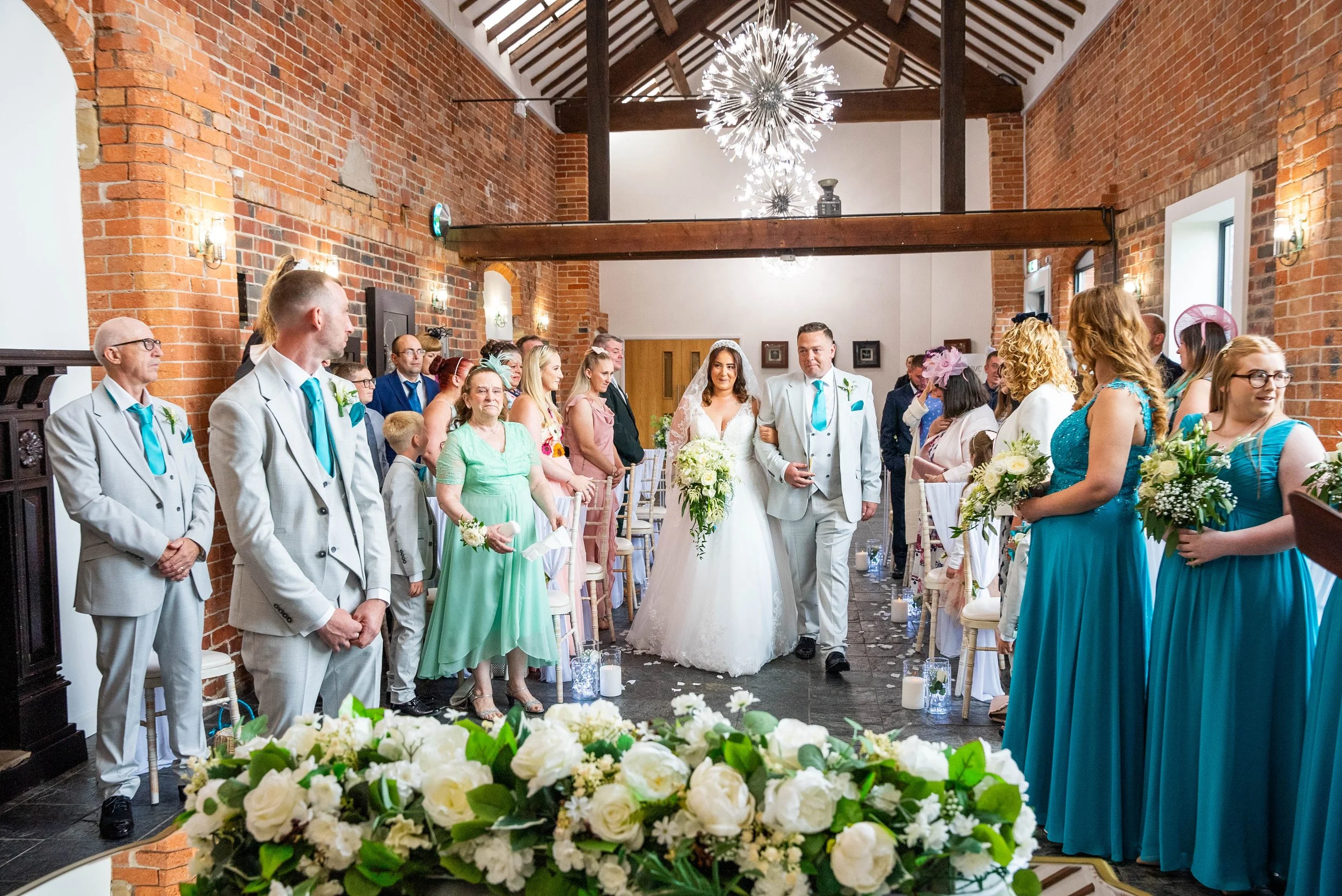 A bride and groom walking down the aisle at a wedding ceremony in a rustic brick venue, surrounded by bridesmaids and groomsmen.