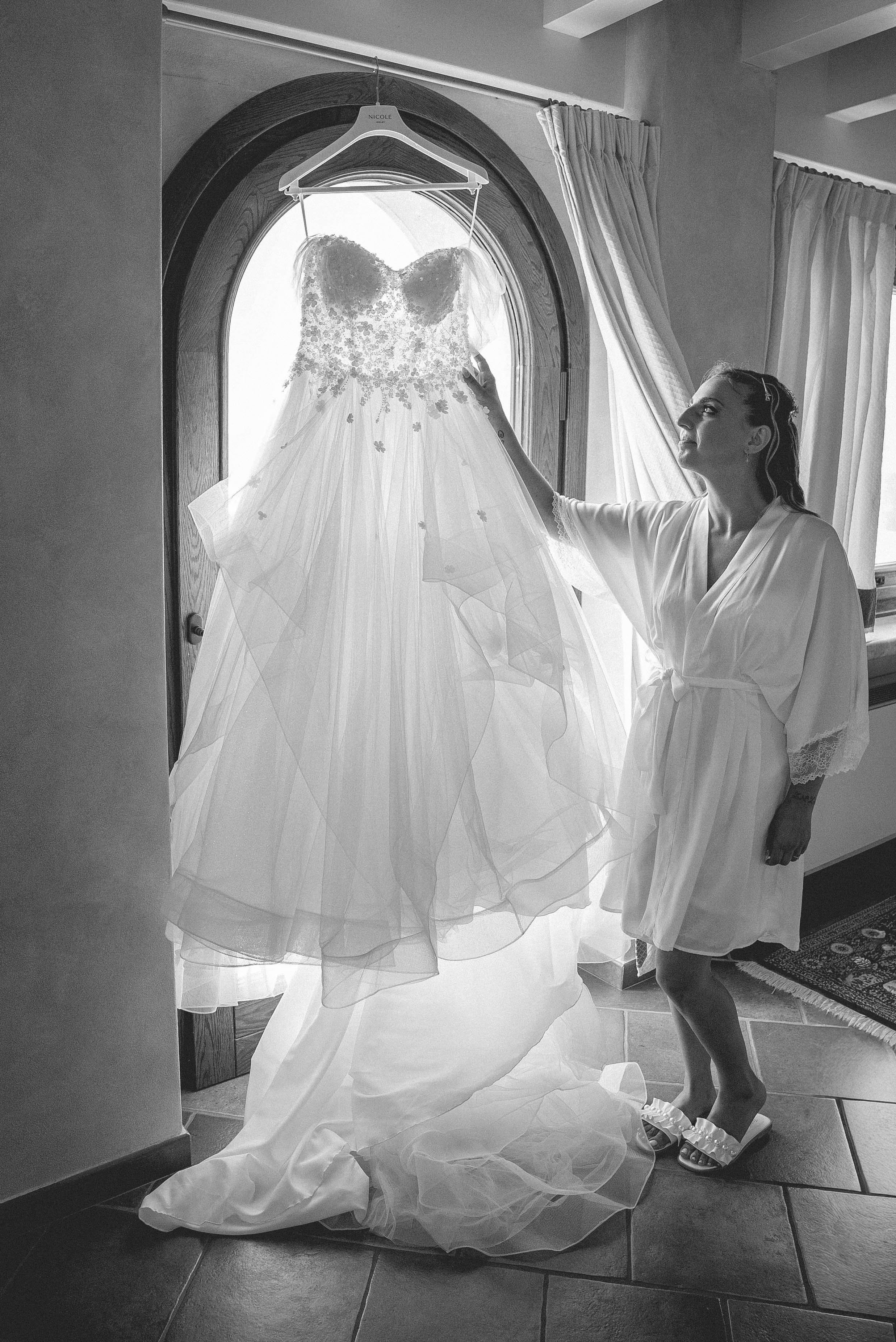 A woman in a white robe is looking at a wedding dress hanging in front of a window. The wedding dress has a floral lace bodice and a full tulle skirt, and it is hanging on a hanger labeled with her name, Nicole. The woman appears to be admiring the d