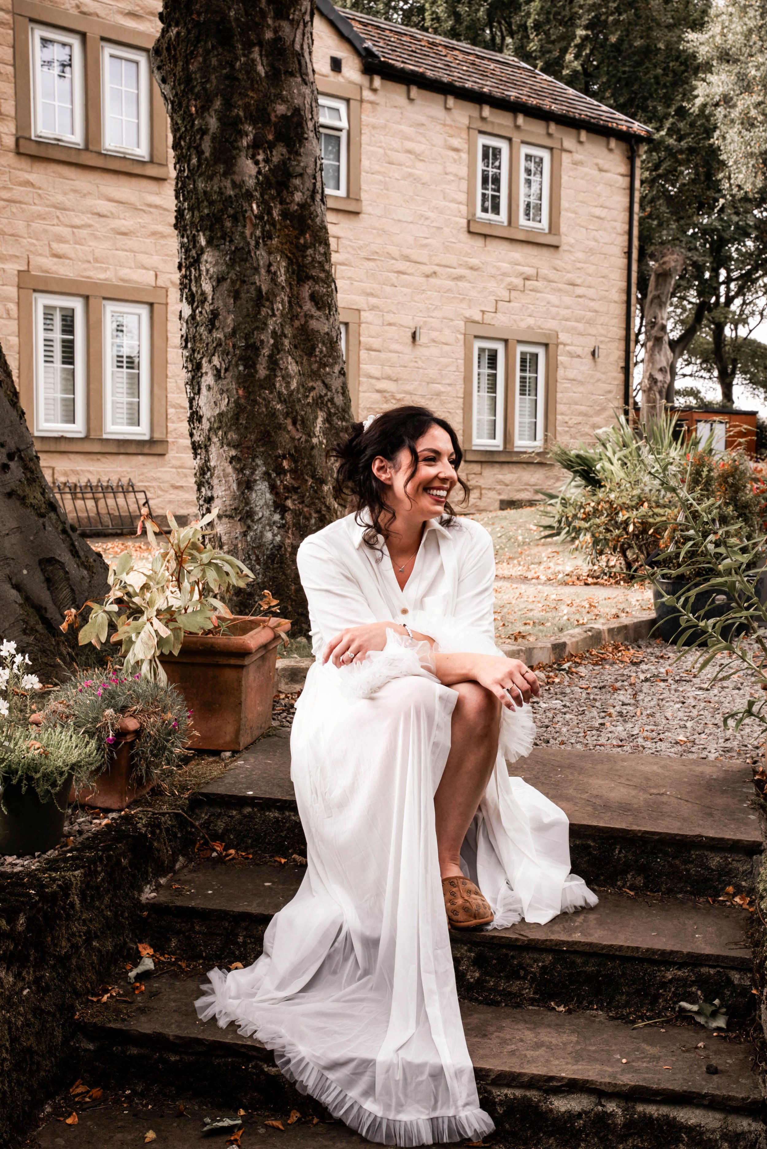 A woman in a white dress sitting on outdoor stone steps, smiling, with a house and trees in the background.