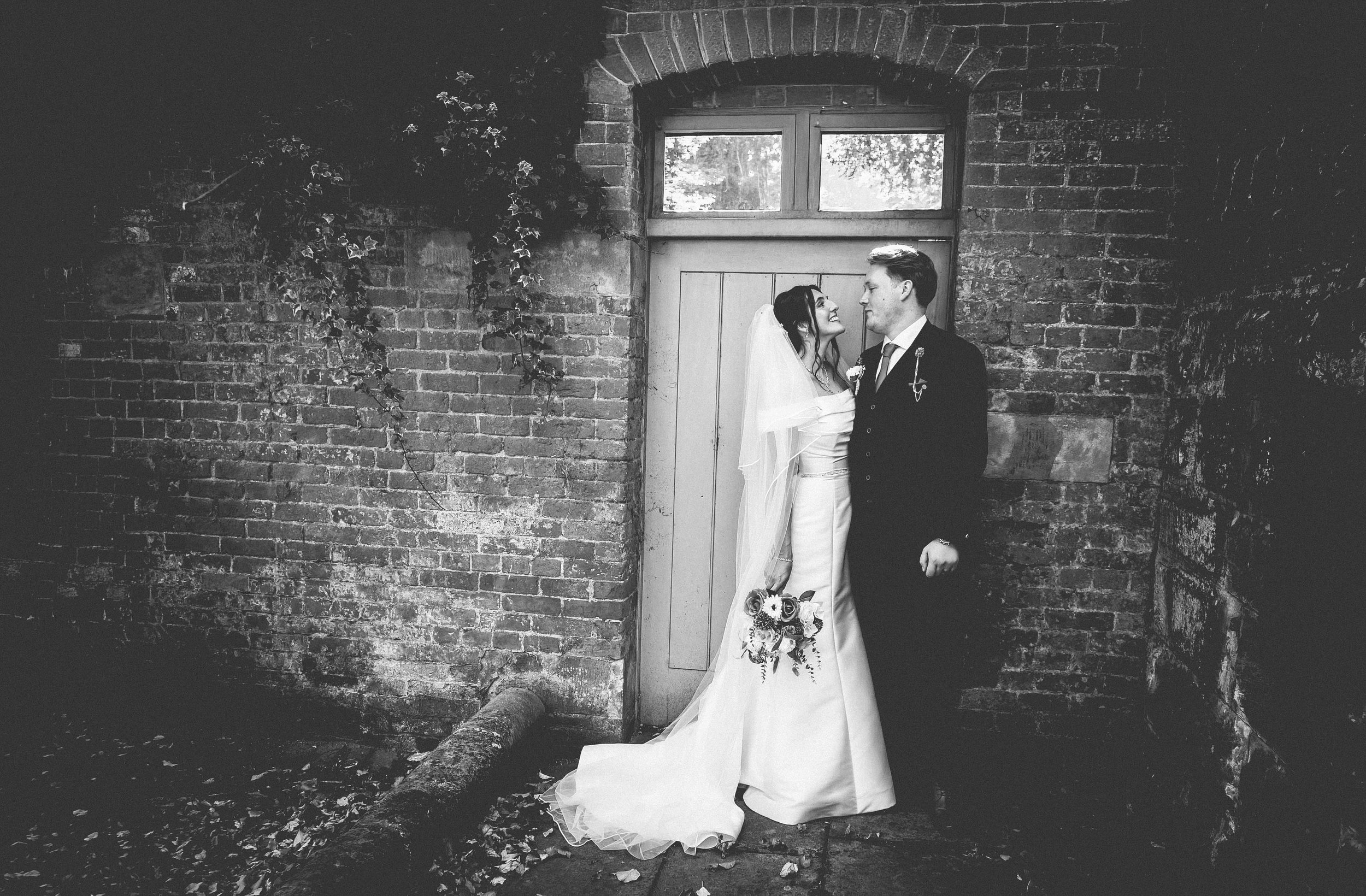 Black and white photo of a bride and groom standing close together in front of a brick wall and wooden door, smiling at each other, with the bride holding a bouquet.