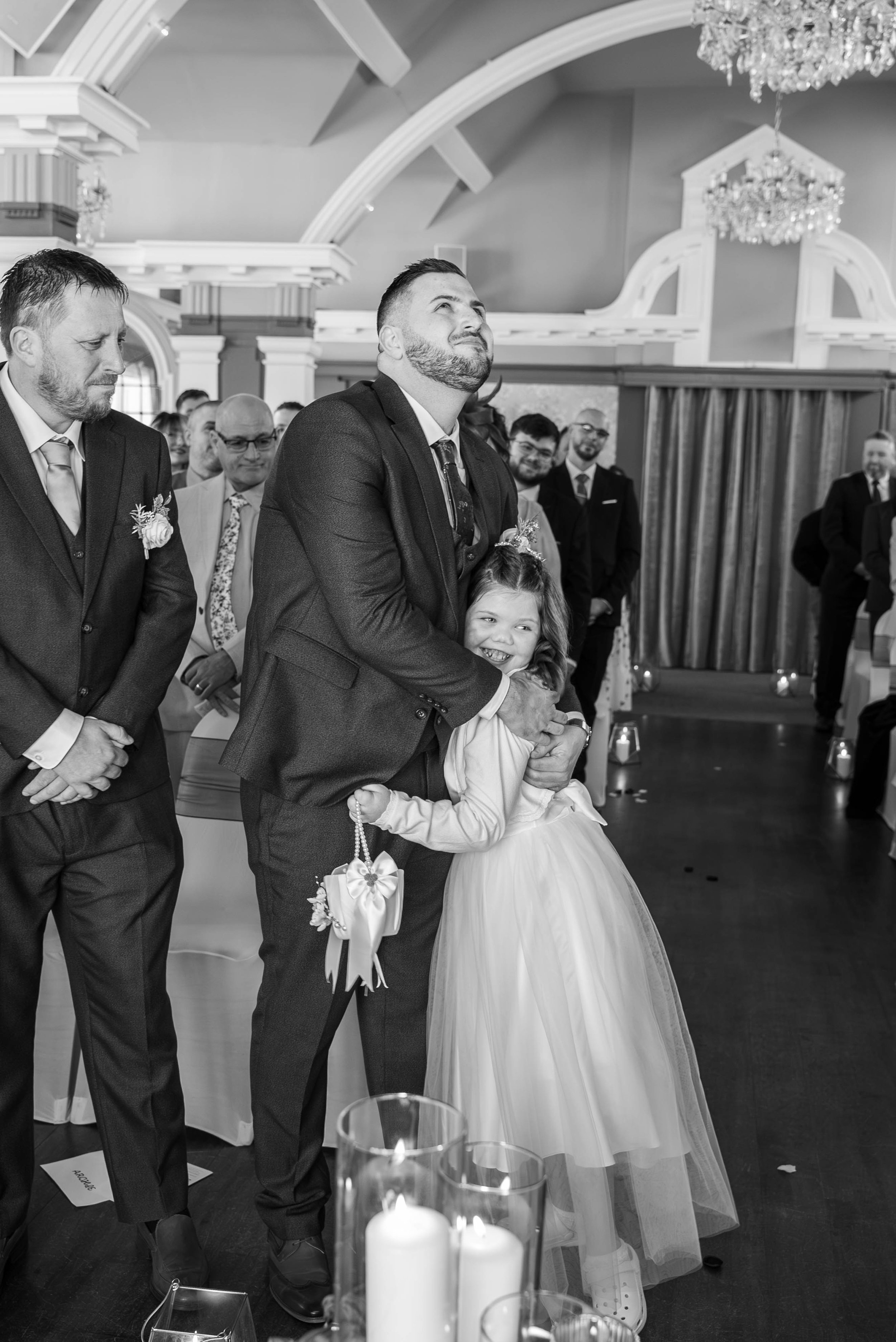 A group of people, including a man and a young girl, standing in a decorated indoor venue during a wedding ceremony. The man is embracing the girl, who is holding a small bag, and the girl is smiling. The scene is in black and white.