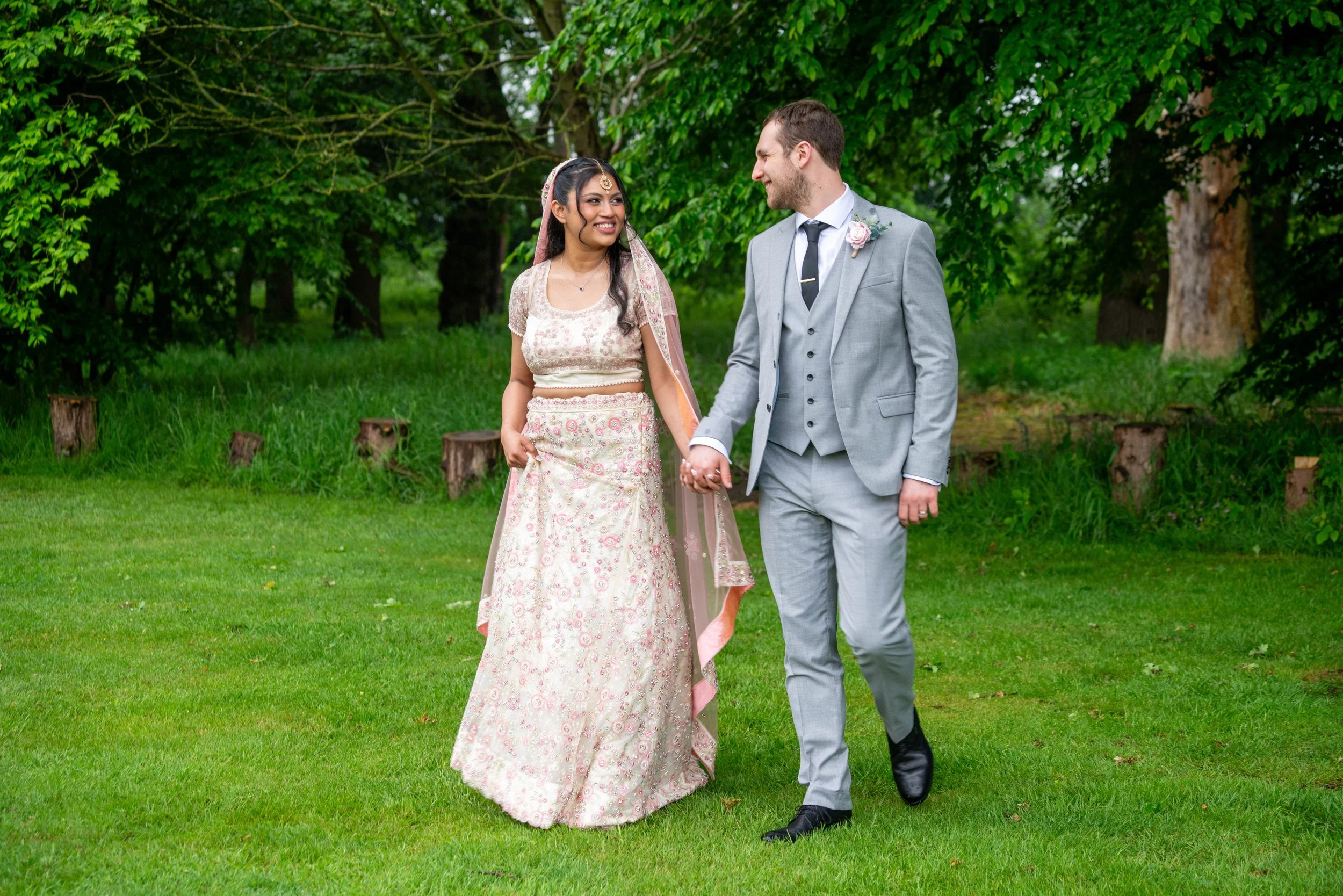 A bride and groom walking hand in hand outdoors on a grassy area, surrounded by green trees.