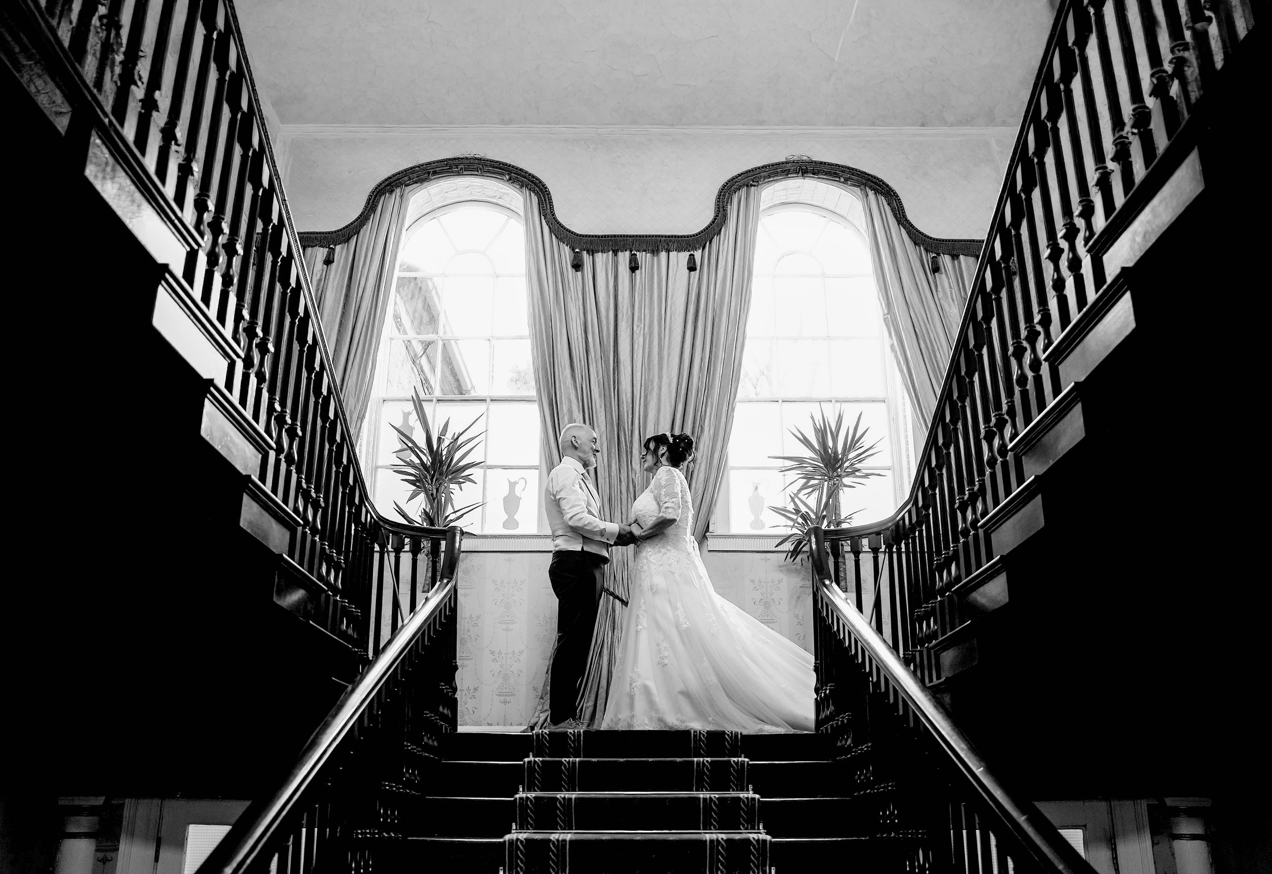 A black and white photo of a bride and groom standing face-to-face on a staircase inside a large, elegant building with high windows, draped curtains, and potted plants, holding hands and looking at each other.