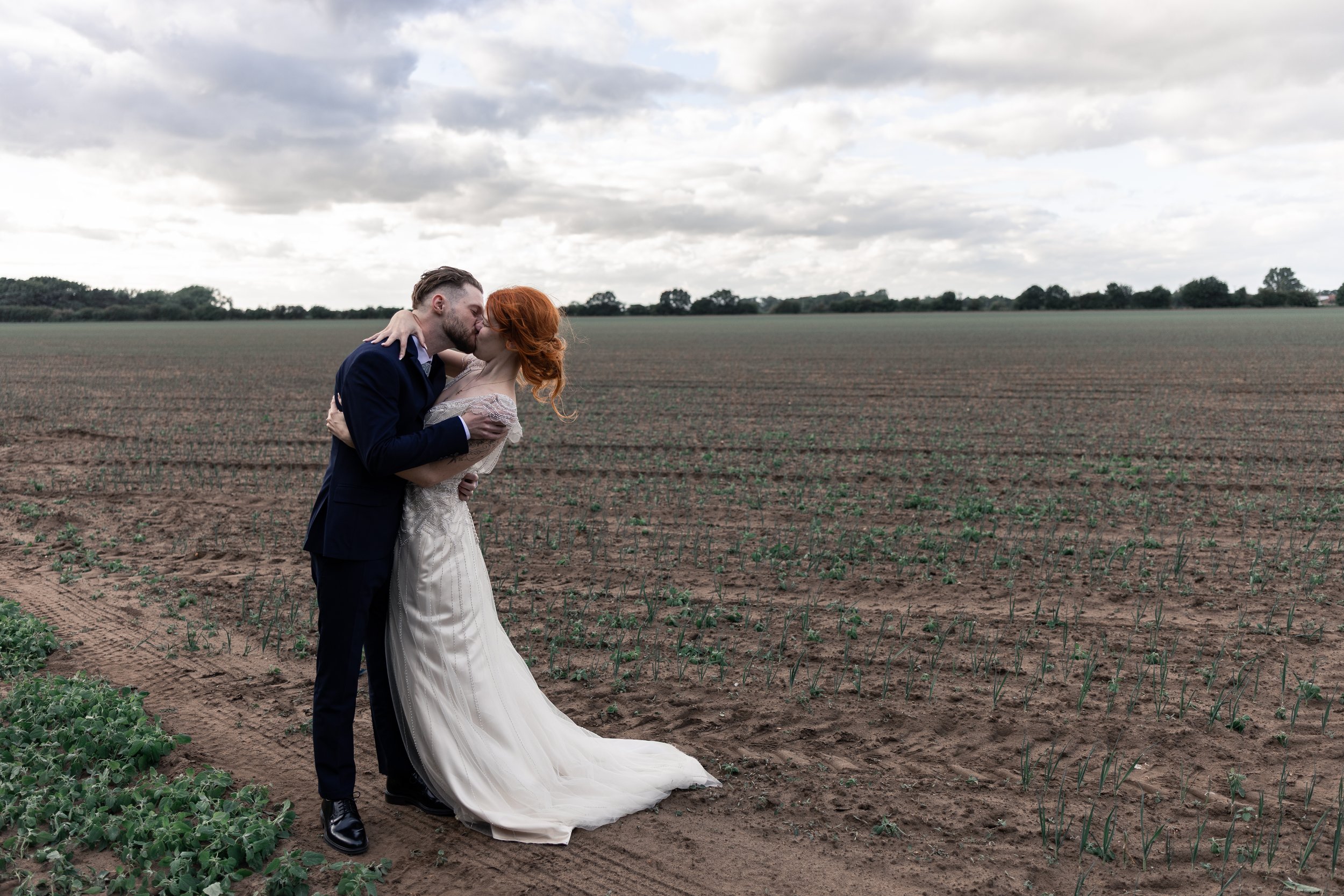 A couple wearing wedding attire, the man in a navy suit and the woman in a white wedding dress, sharing a kiss in a farmland field under a cloudy sky.