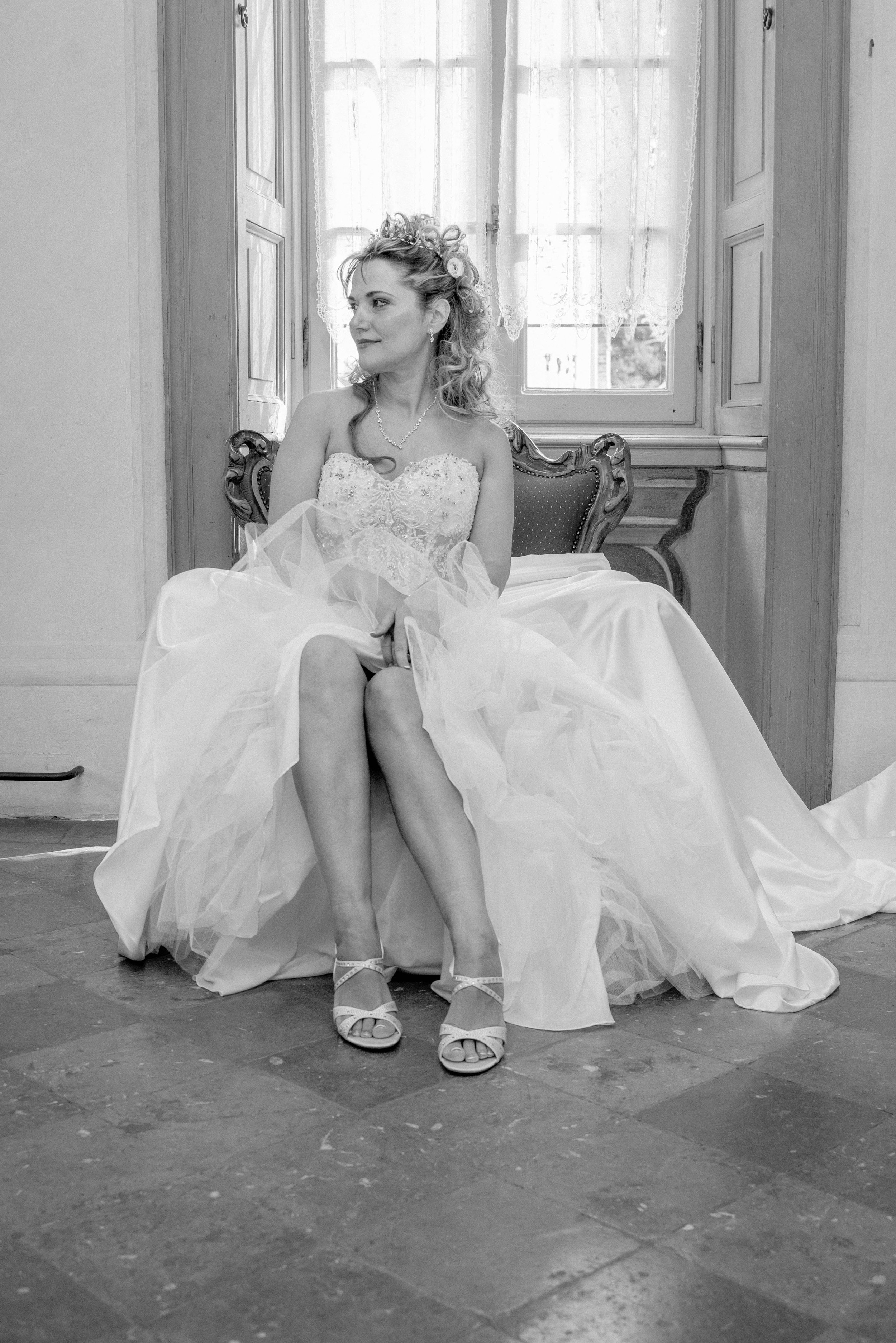 A woman in a wedding dress sitting on a vintage chair in a room with window curtains.