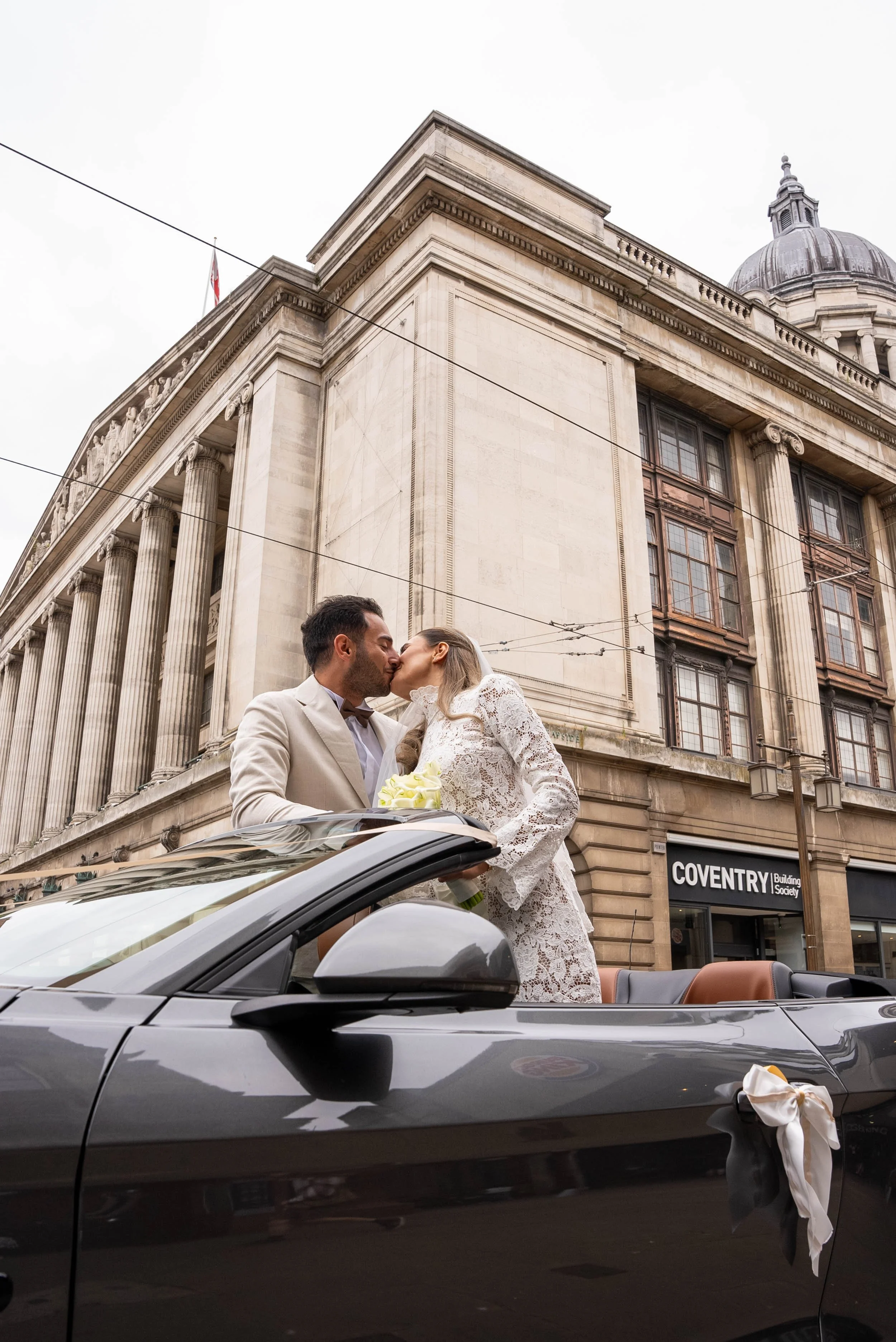 A newlywed couple sharing a kiss inside a convertible car, with a historic building in the background.
